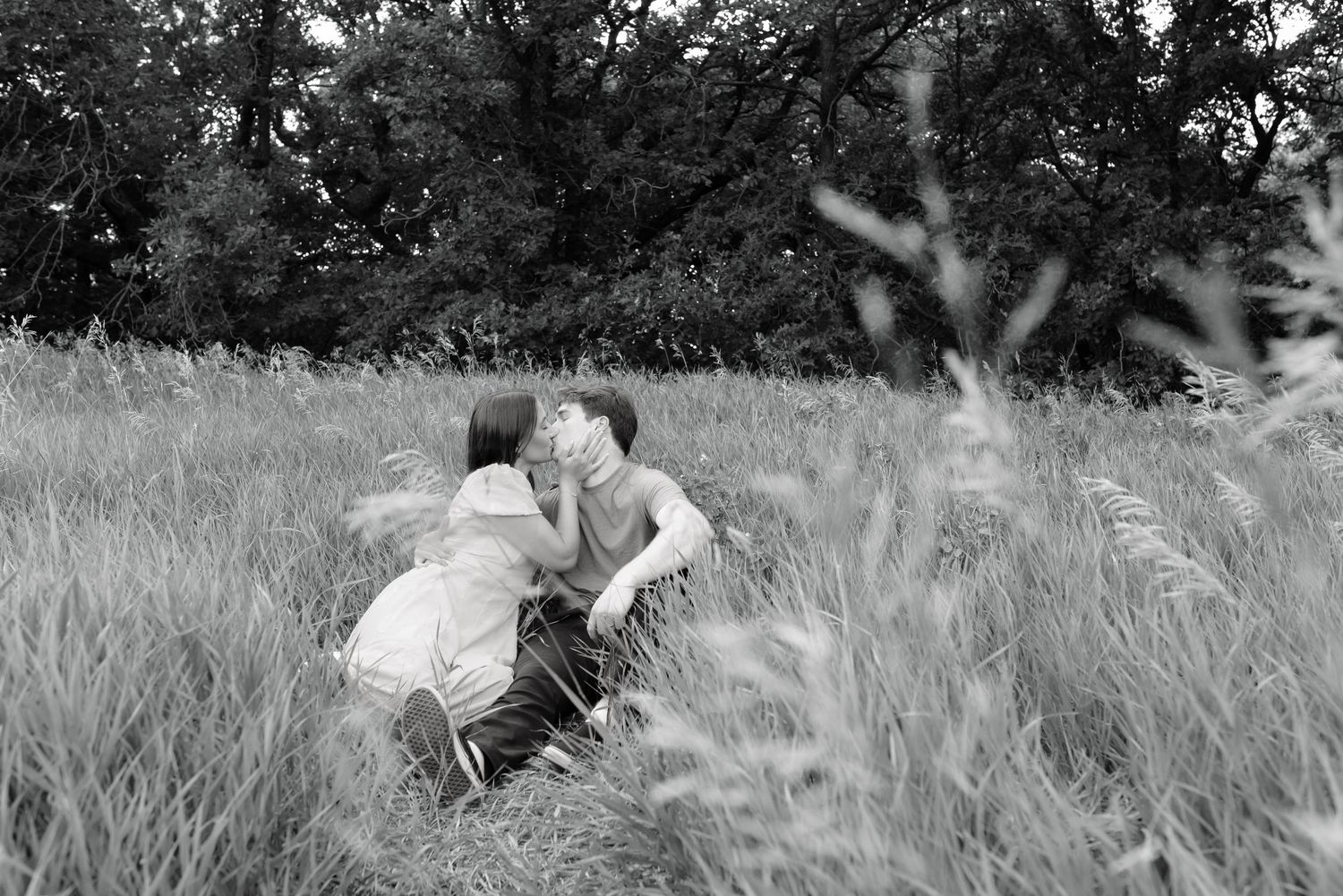 Black and white photo of a couple sharing a kiss in a tall grass field with trees in the background.