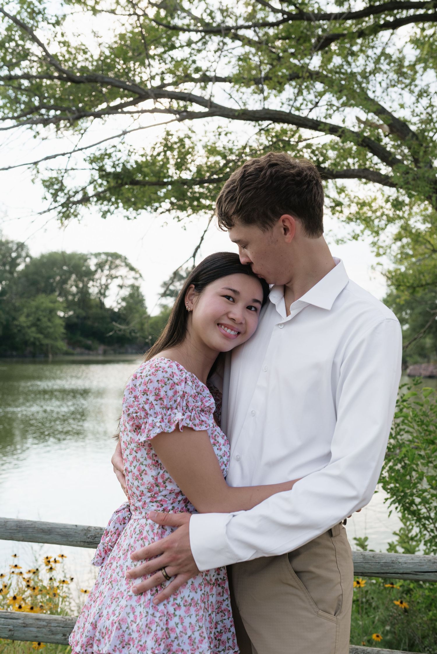 A couple embracing near a wooden fence by a tranquil lake, woman smiling in a floral dress.