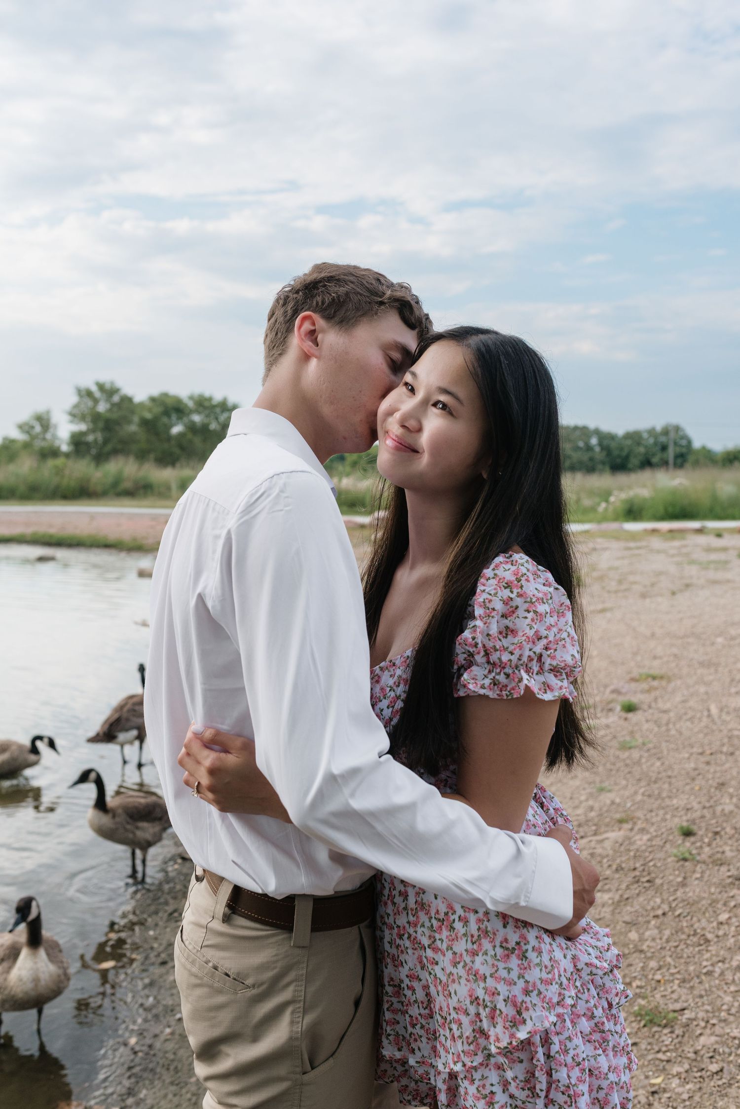 A couple embraces near a lakeside shore with Canada geese wading in the water behind them.