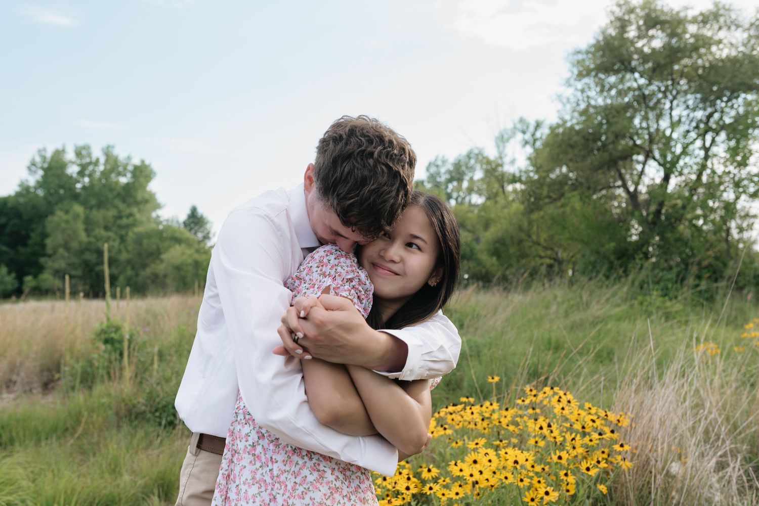 Man in white shirt hugging woman in floral dress near yellow wildflowers in a meadow.