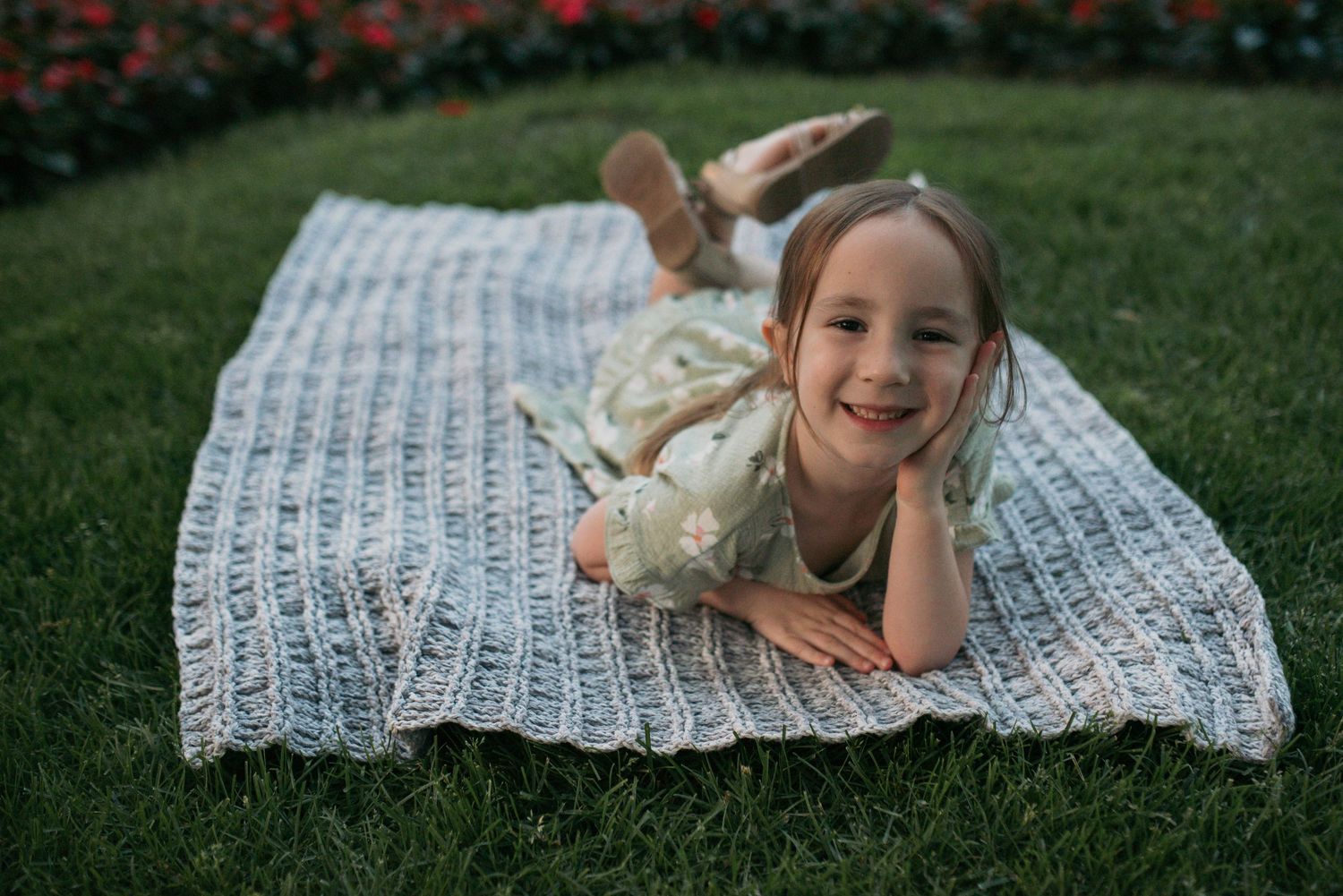 A smiling young girl in a floral dress lying on a knit blanket on green grass outdoors.