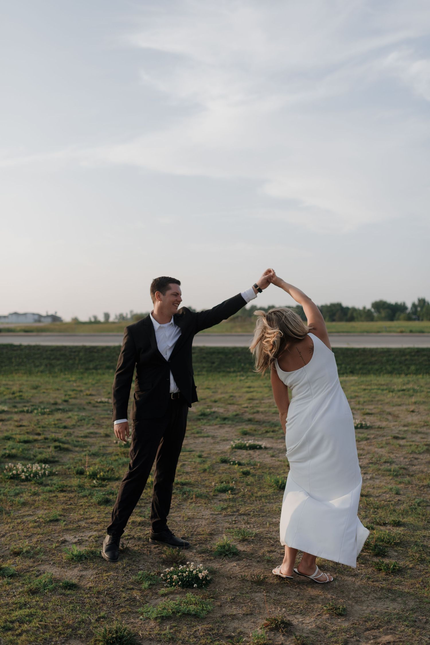 Couple dancing outdoors at sunset, man twirling woman in elegant white dress in grassy field.