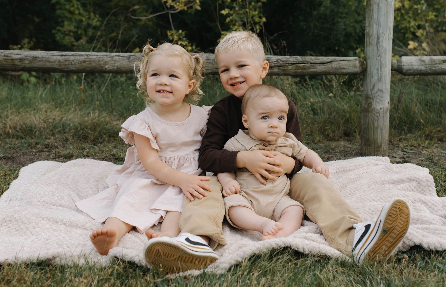 Three young siblings sitting on a blanket outdoors, two toddlers and a baby posing for a family portrait.