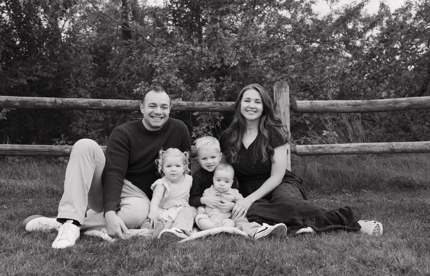 Family of five with two parents and three young children sit together on grass in black and white.