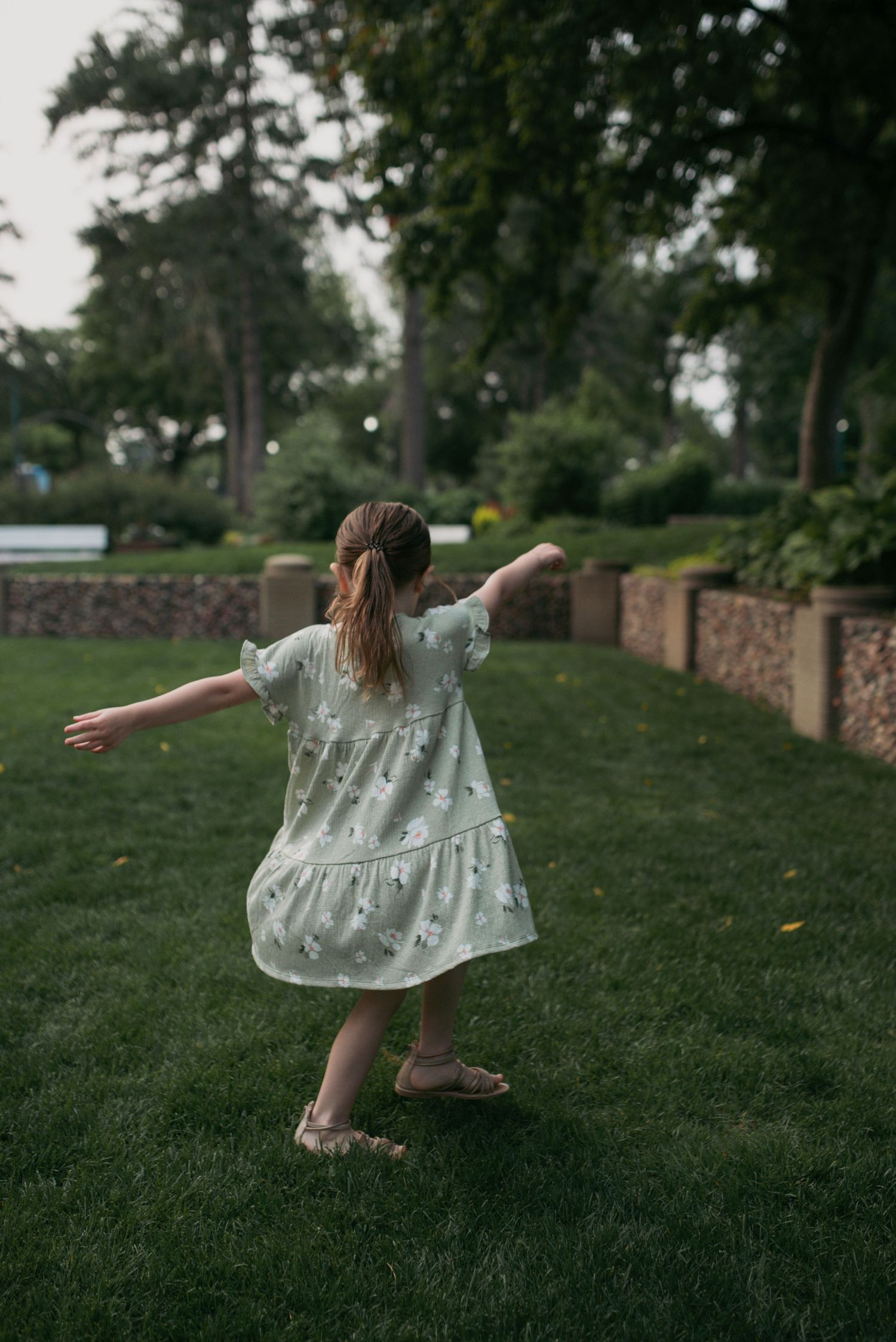 Child in mint floral dress dancing with arms outstretched on green lawn in a park.