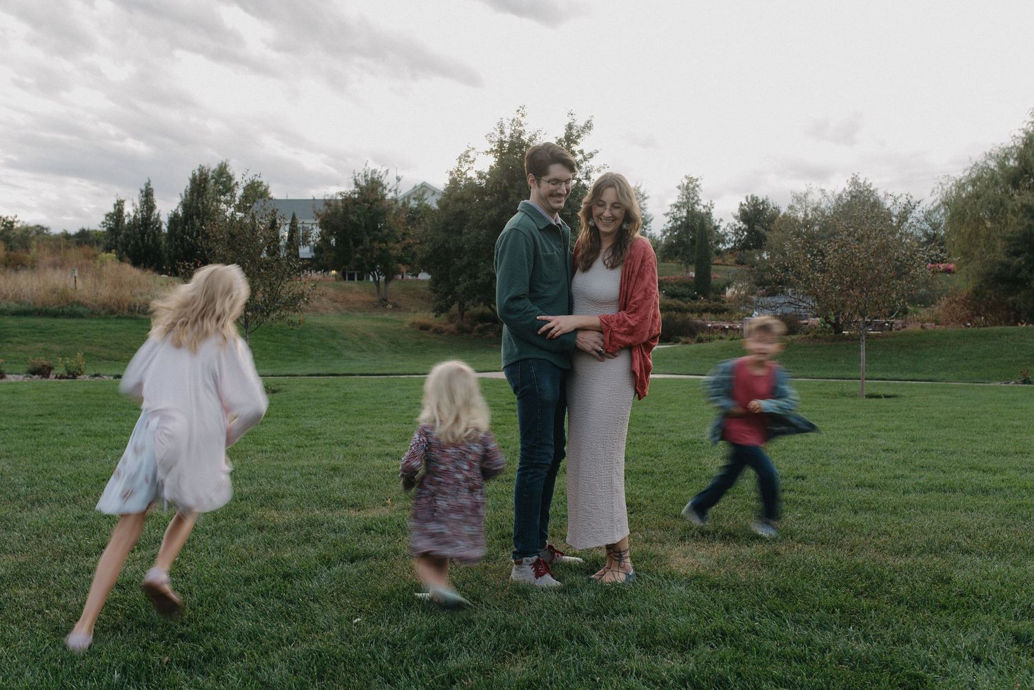 Parents standing together on a lawn while three children play around them.