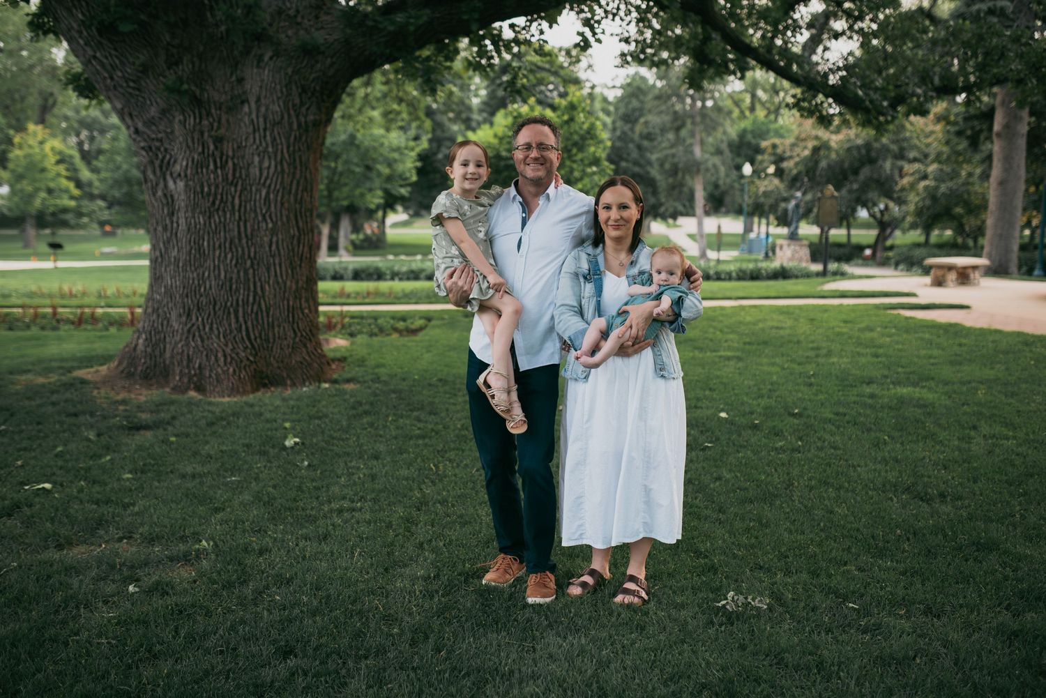 Happy family of four posing outdoors in a green park, parents holding two young children.
