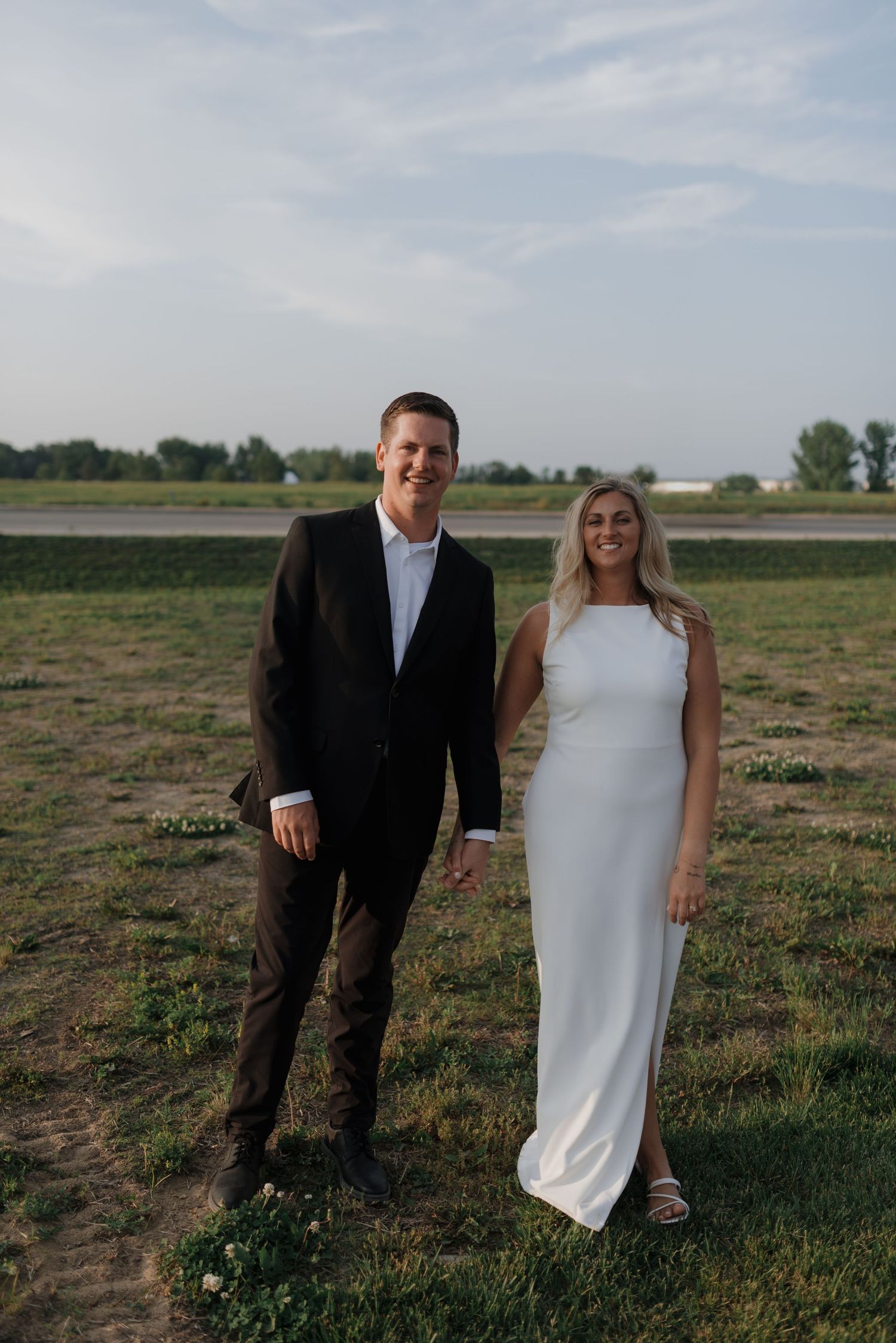 Smiling bride in white gown and groom in black suit holding hands outdoors in a grassy field.