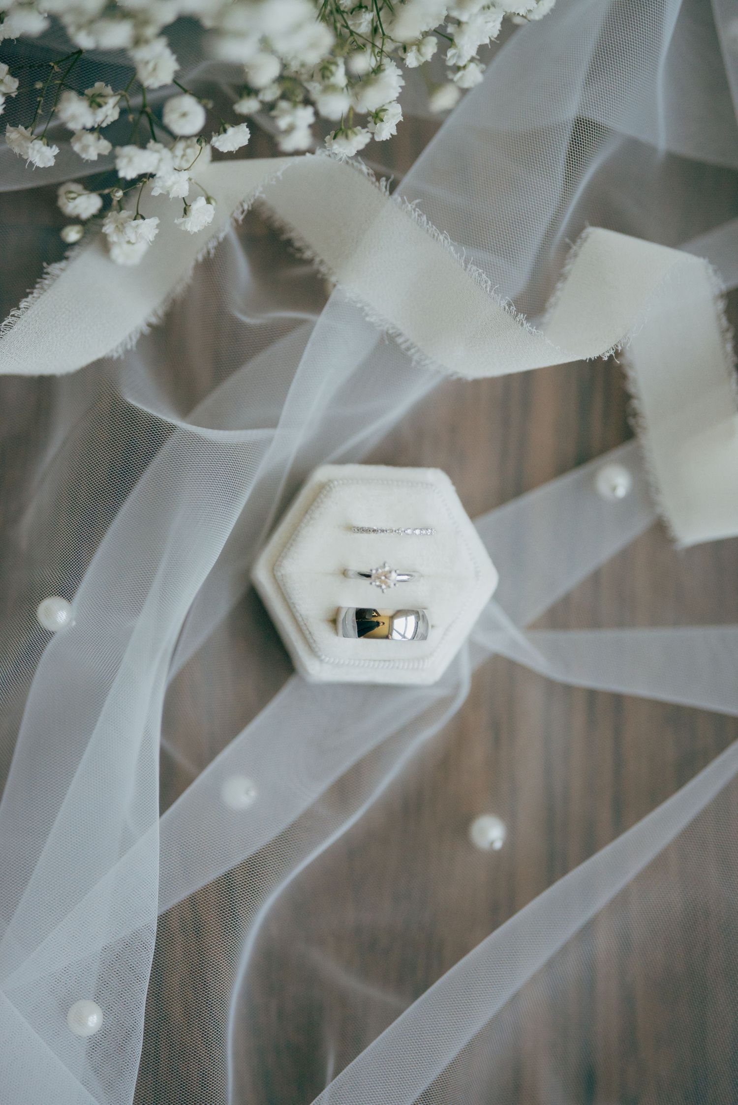 Wedding rings in a hexagonal velvet box surrounded by a white veil, pearls, and baby's breath flowers on a wood surface.