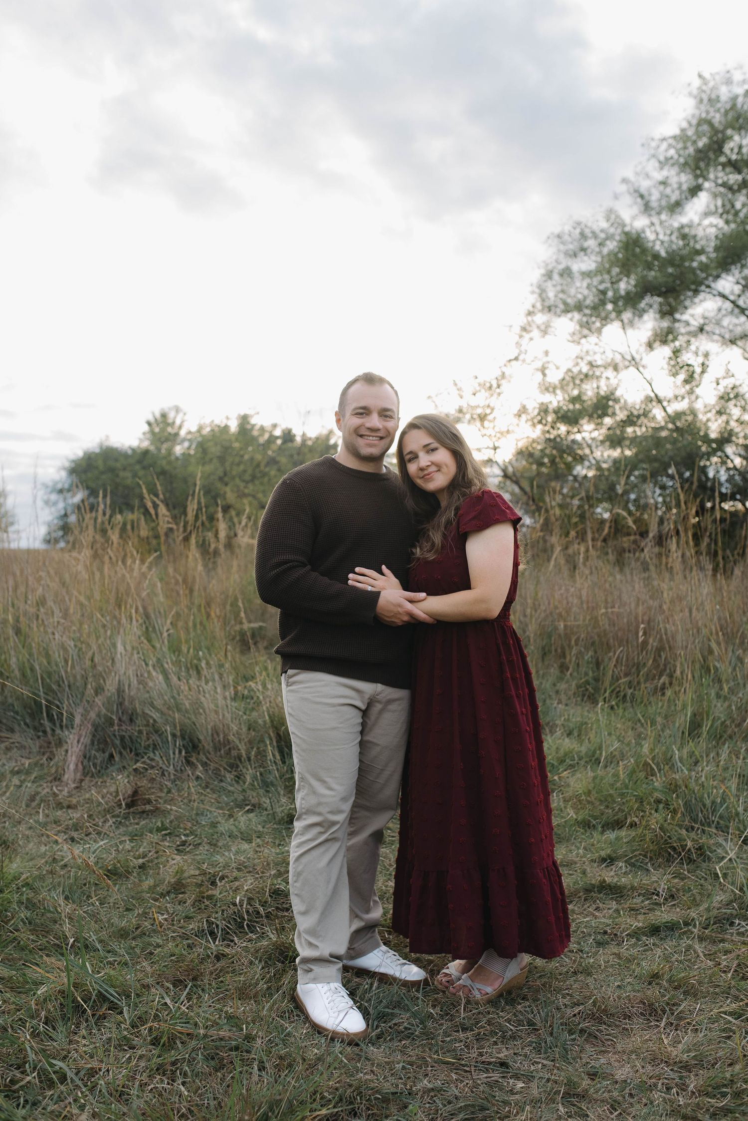 A couple embraces in a golden meadow at sunset, woman in burgundy dress and man in dark sweater.
