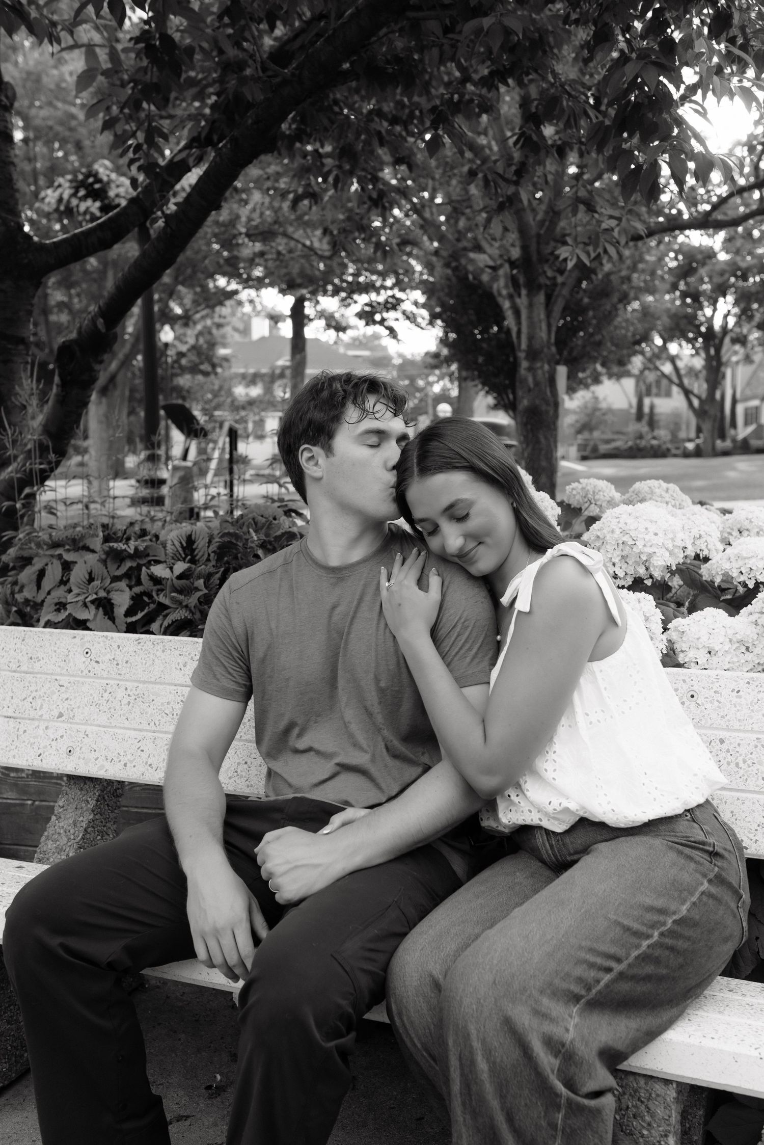 Black and white photo of a couple sitting on a bench, man kissing woman's head as she leans on his shoulder.