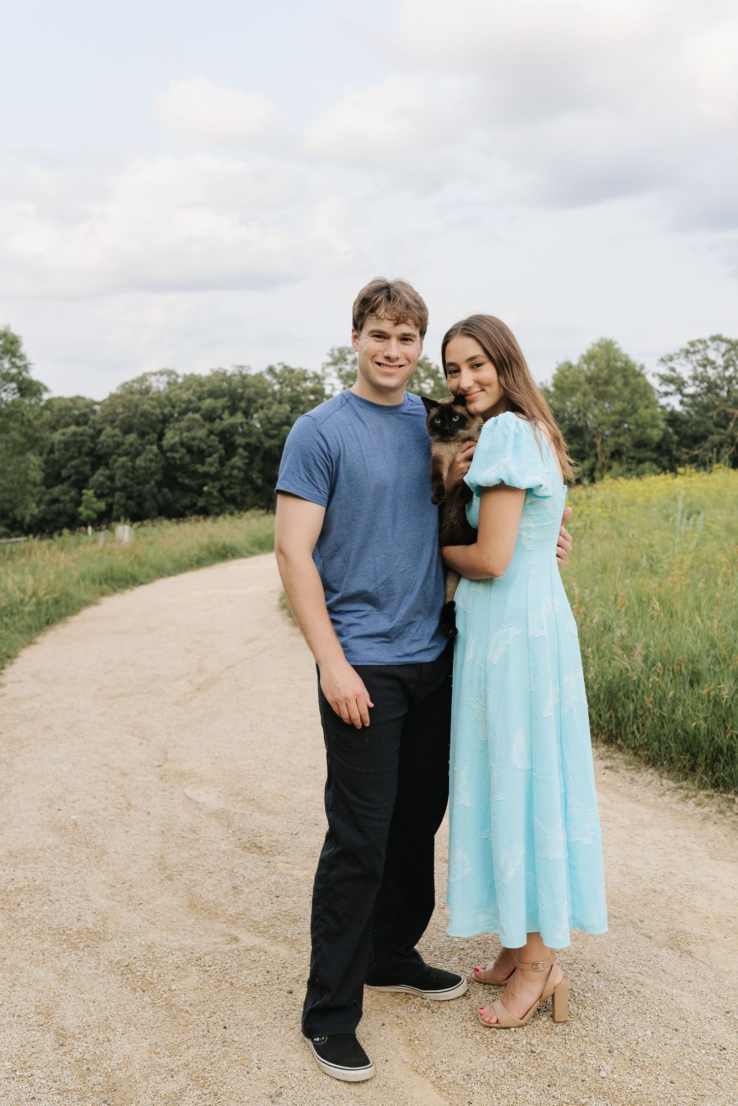 A young couple smiles on a dirt path holding a small dog, with green fields and trees in the background.