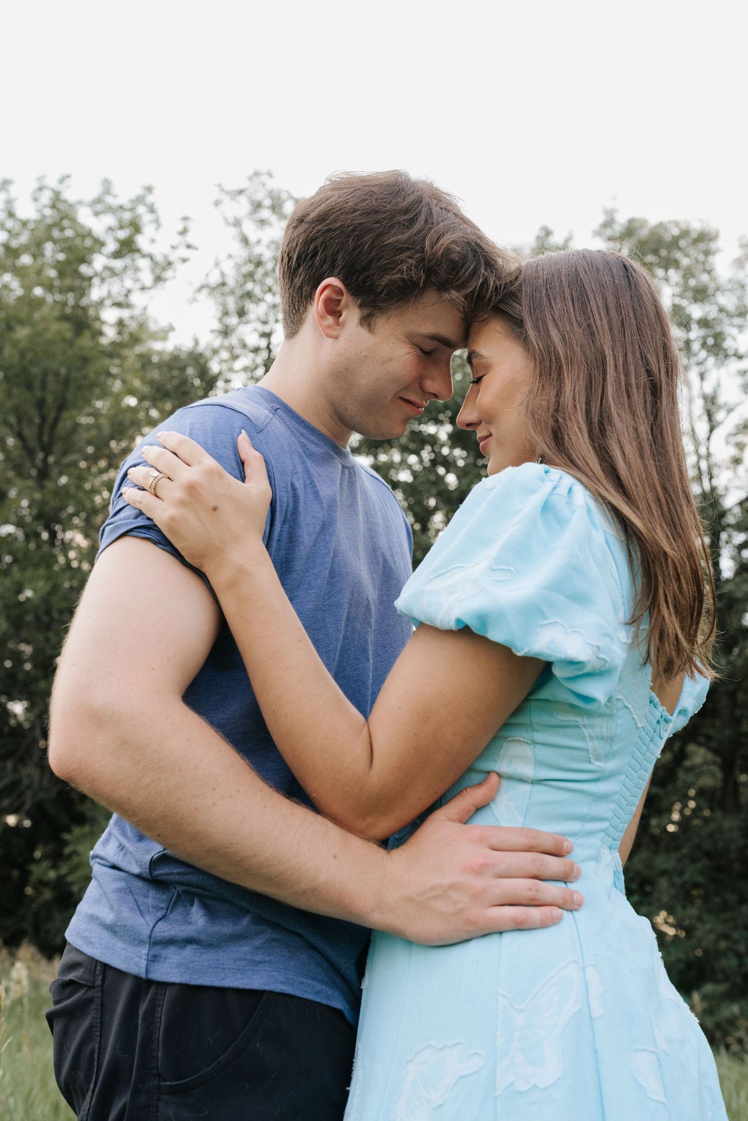 A young couple embracing outdoors, touching foreheads romantically, wearing casual blue outfits with green trees behind them.