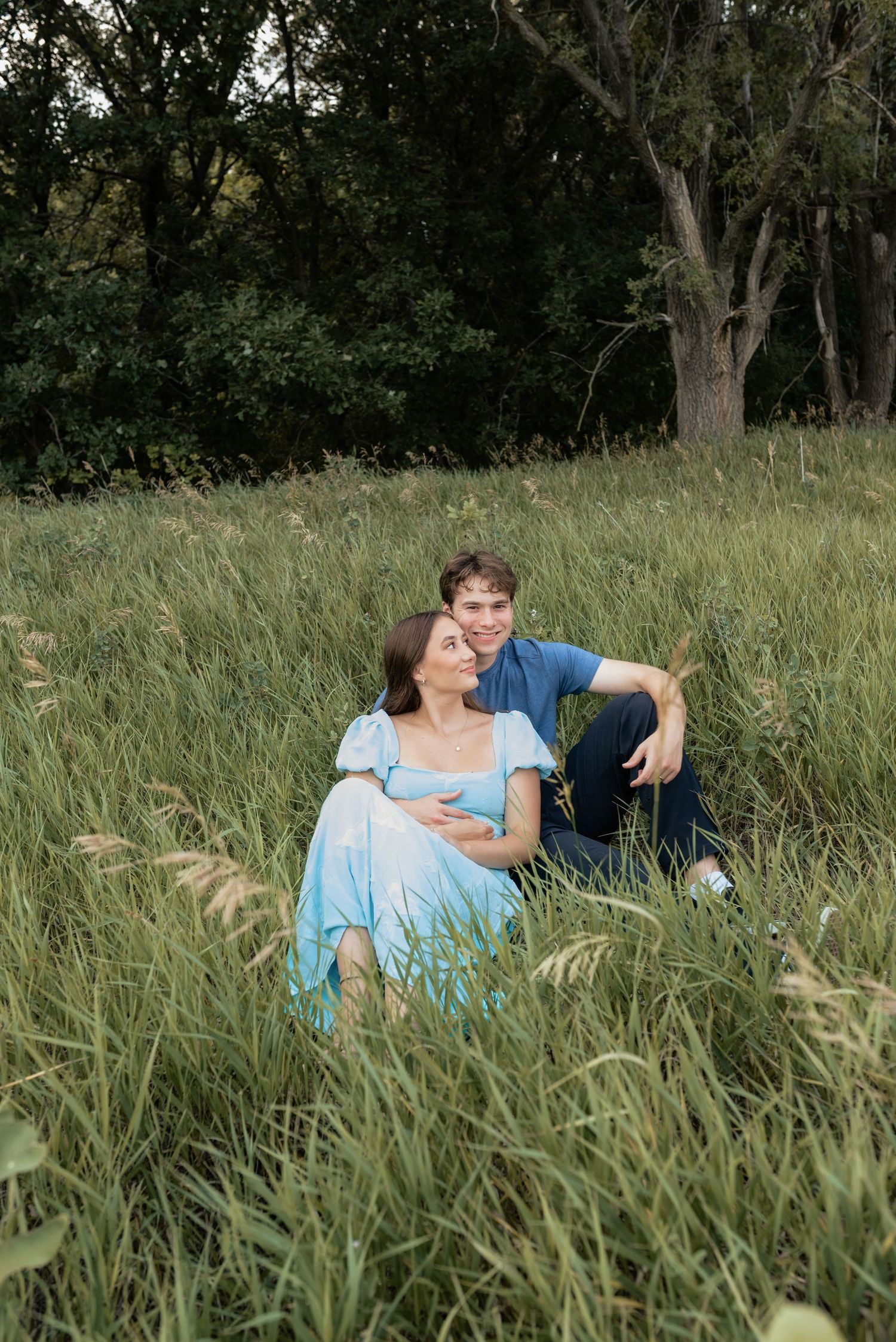 A couple sits together in tall green grass, the woman in a blue dress leaning against the smiling man near trees.