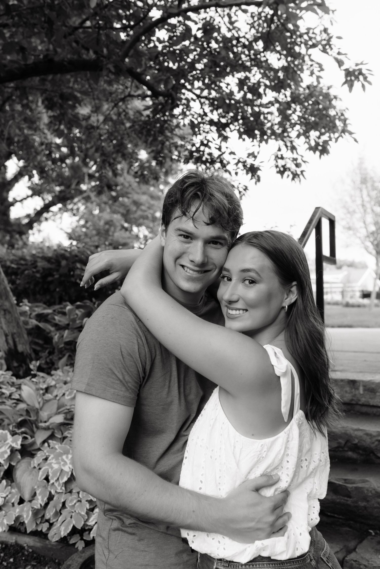 Smiling young couple hugging outdoors in black and white photo, woman with arms around mans neck near garden steps.