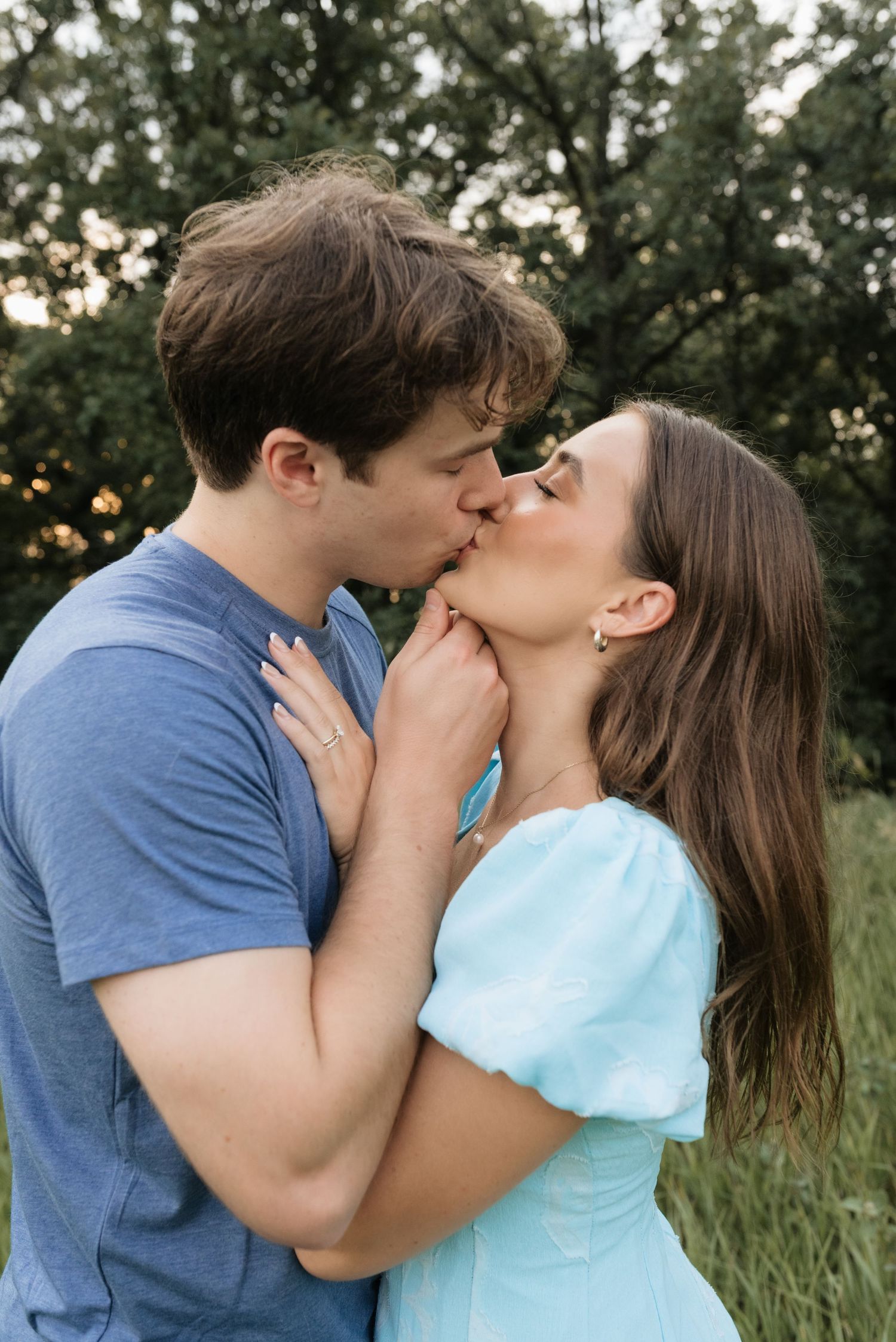 A couple shares a romantic kiss outdoors in a lush green field, man in blue tee and woman in light blue dress.