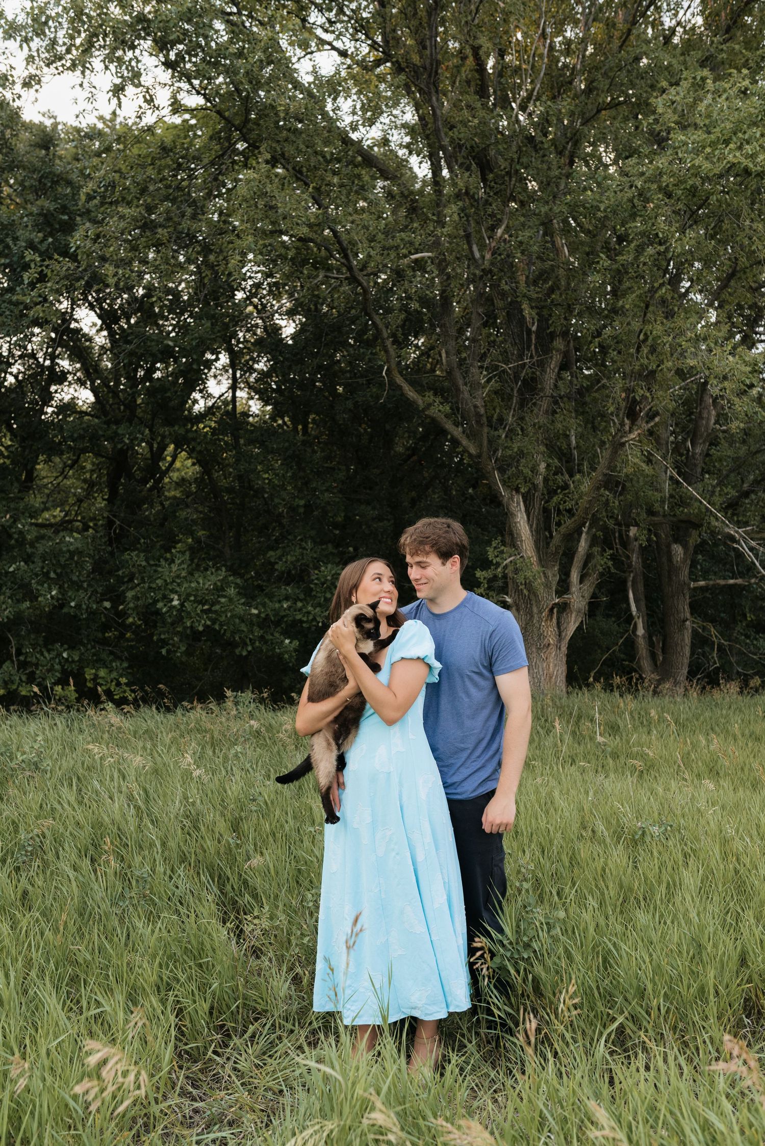 A couple stands in a lush green field, woman in blue dress holding a cat, man in blue t-shirt beside her.