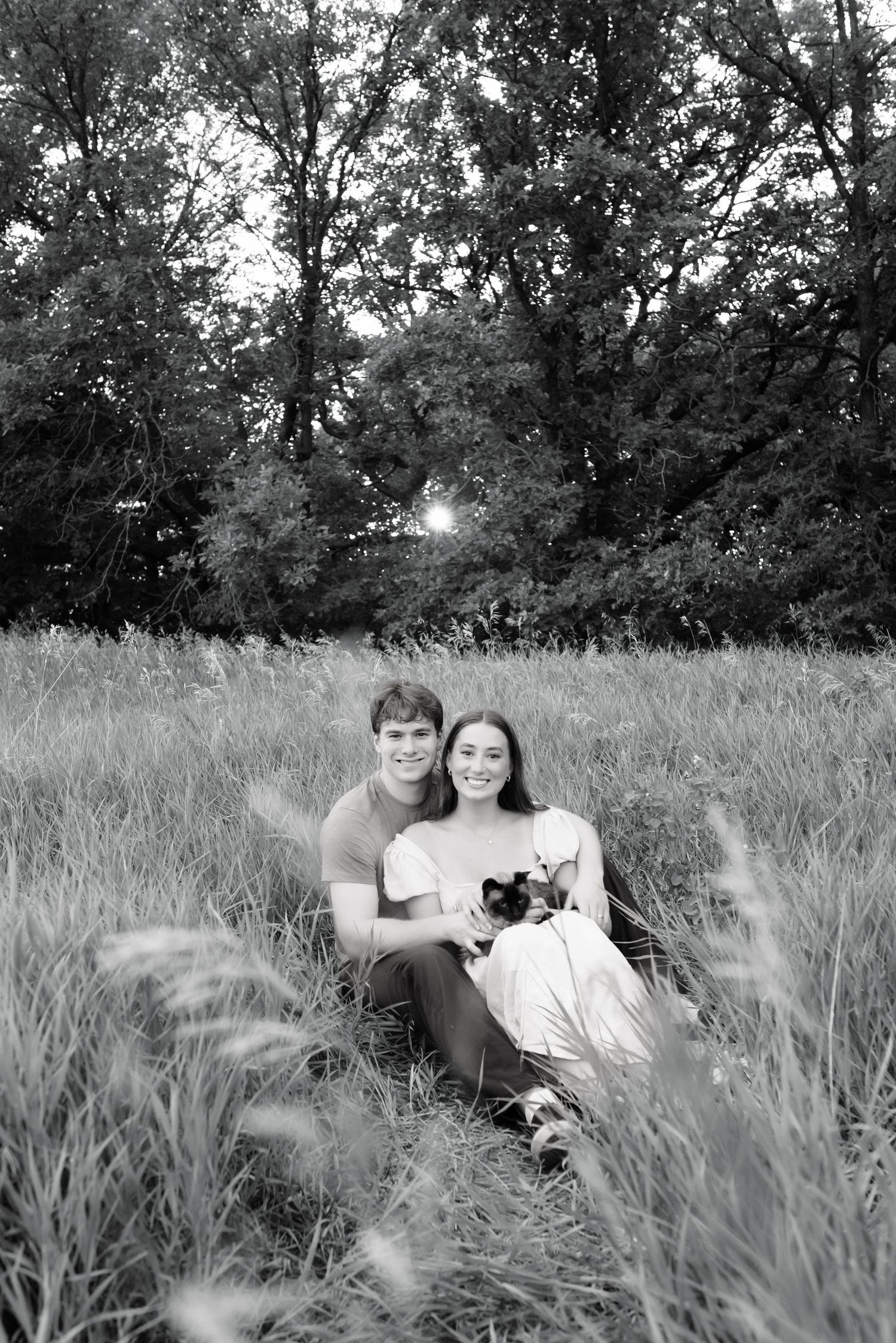 Black and white photo of a smiling couple sitting in a grassy field with trees, holding a small dog.