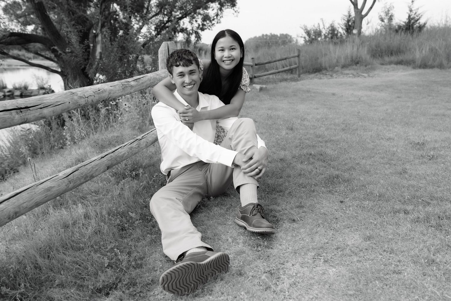 A couple poses playfully by a wooden fence outdoors in a black and white photo.