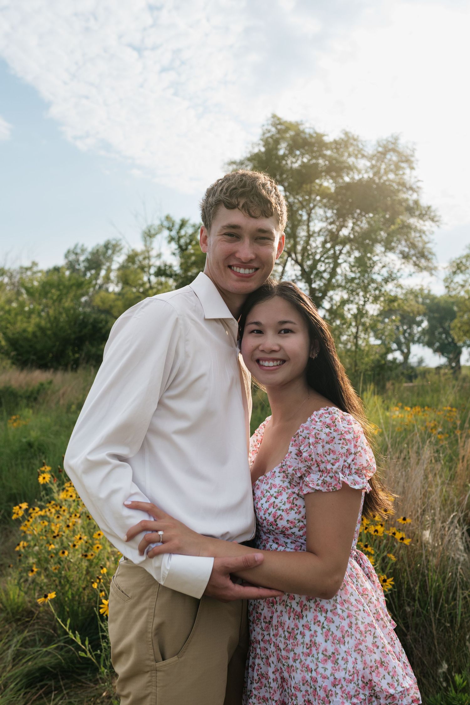 A happy couple hugs and smiles together near yellow wildflowers in a sunlit green meadow.
