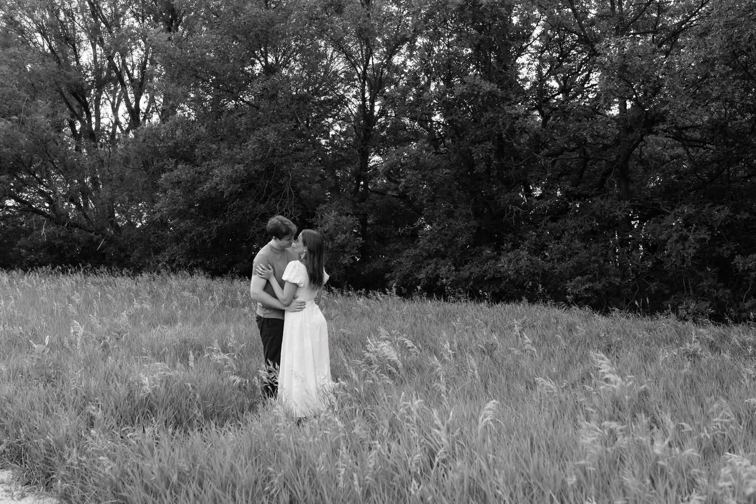 Black and white photo of a couple embracing in a lush green meadow surrounded by trees.
