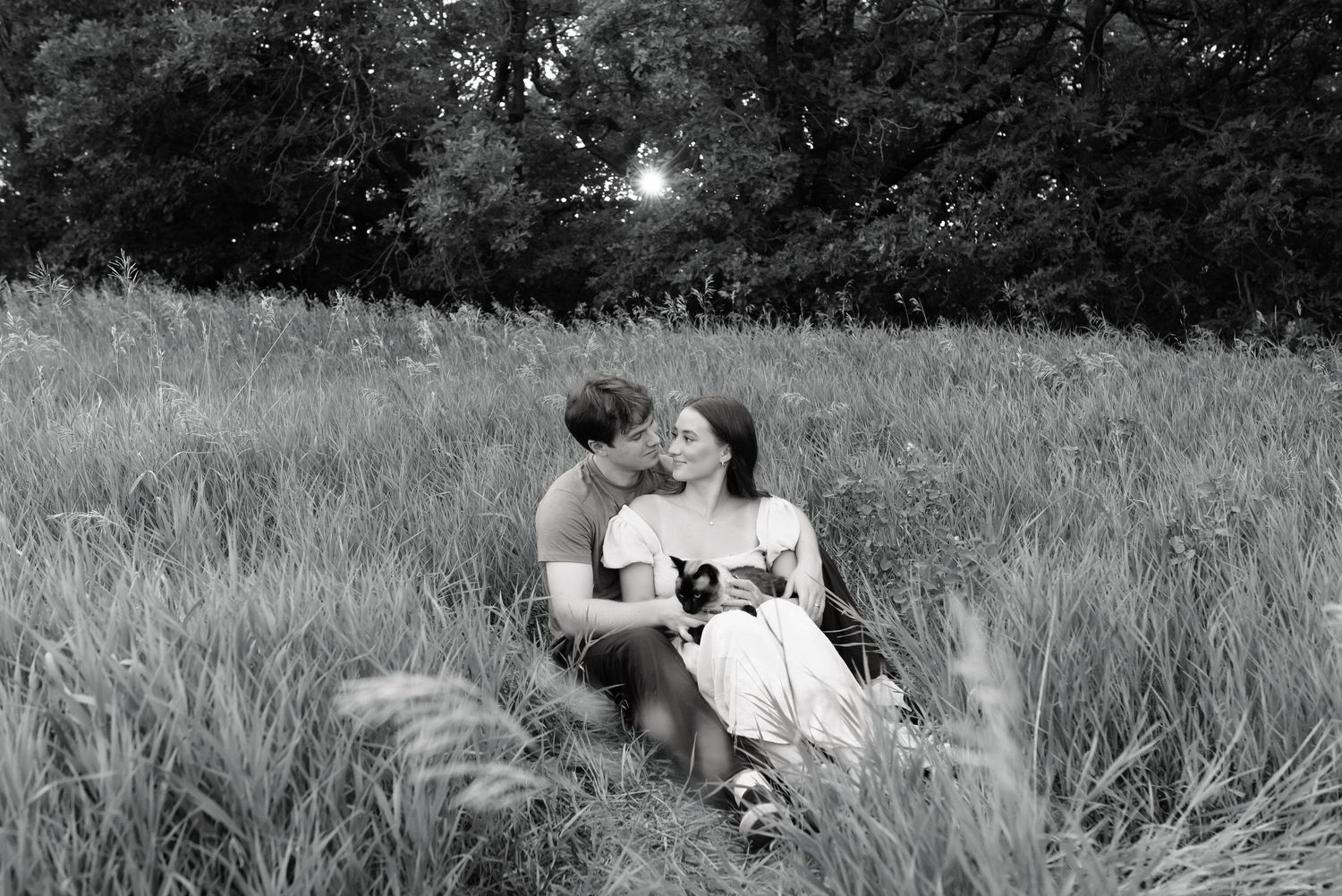 Black and white photo of a romantic couple sitting together in a lush meadow, sharing a tender moment.