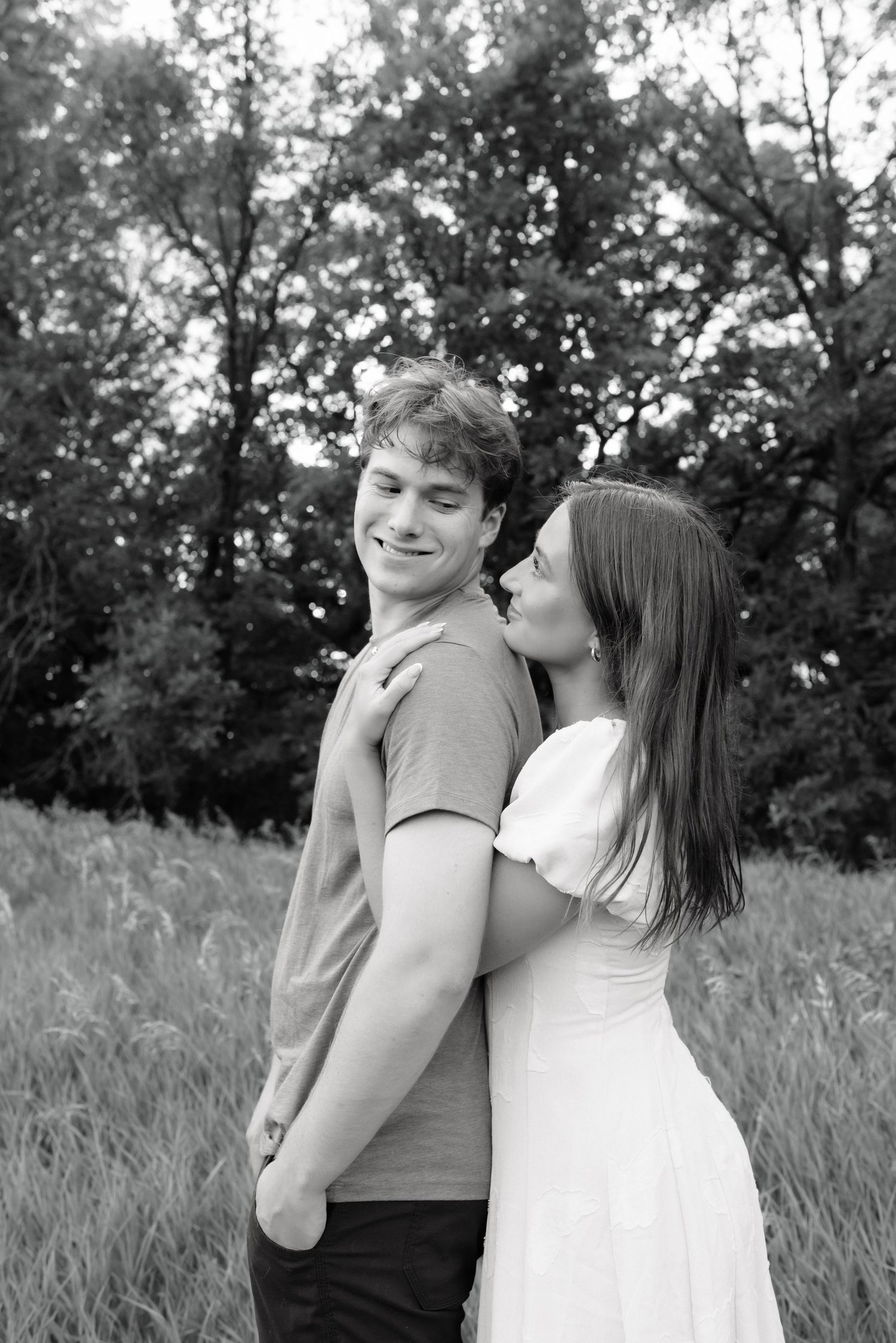 Black and white photo of a young couple embracing outdoors, woman kissing man's cheek in a field.