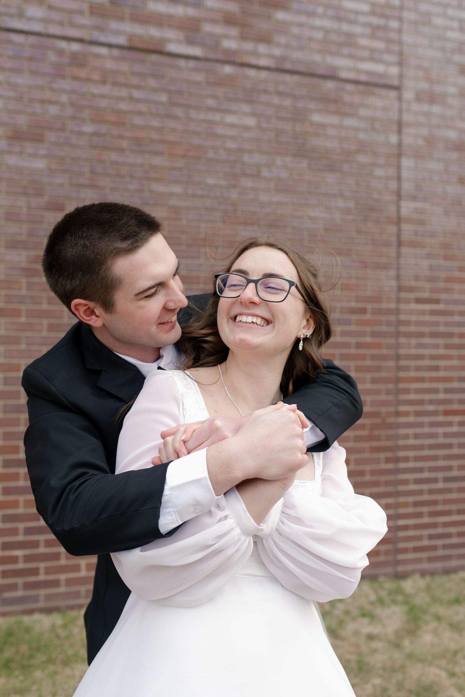 Joyful bride in white gown laughs as groom embraces her from behind against brick wall backdrop.