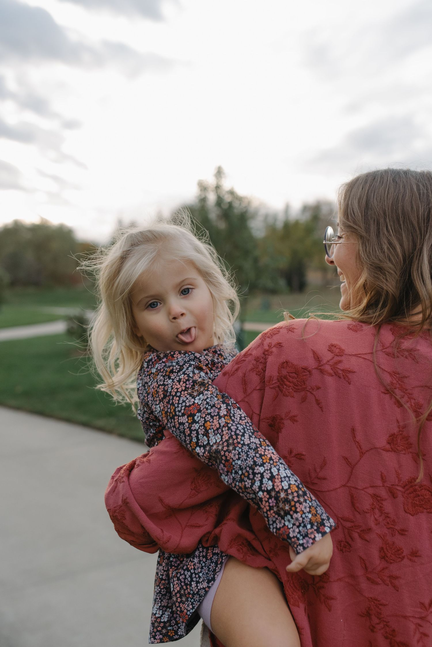 A blonde toddler sticks out her tongue while being carried by a woman in a red top.