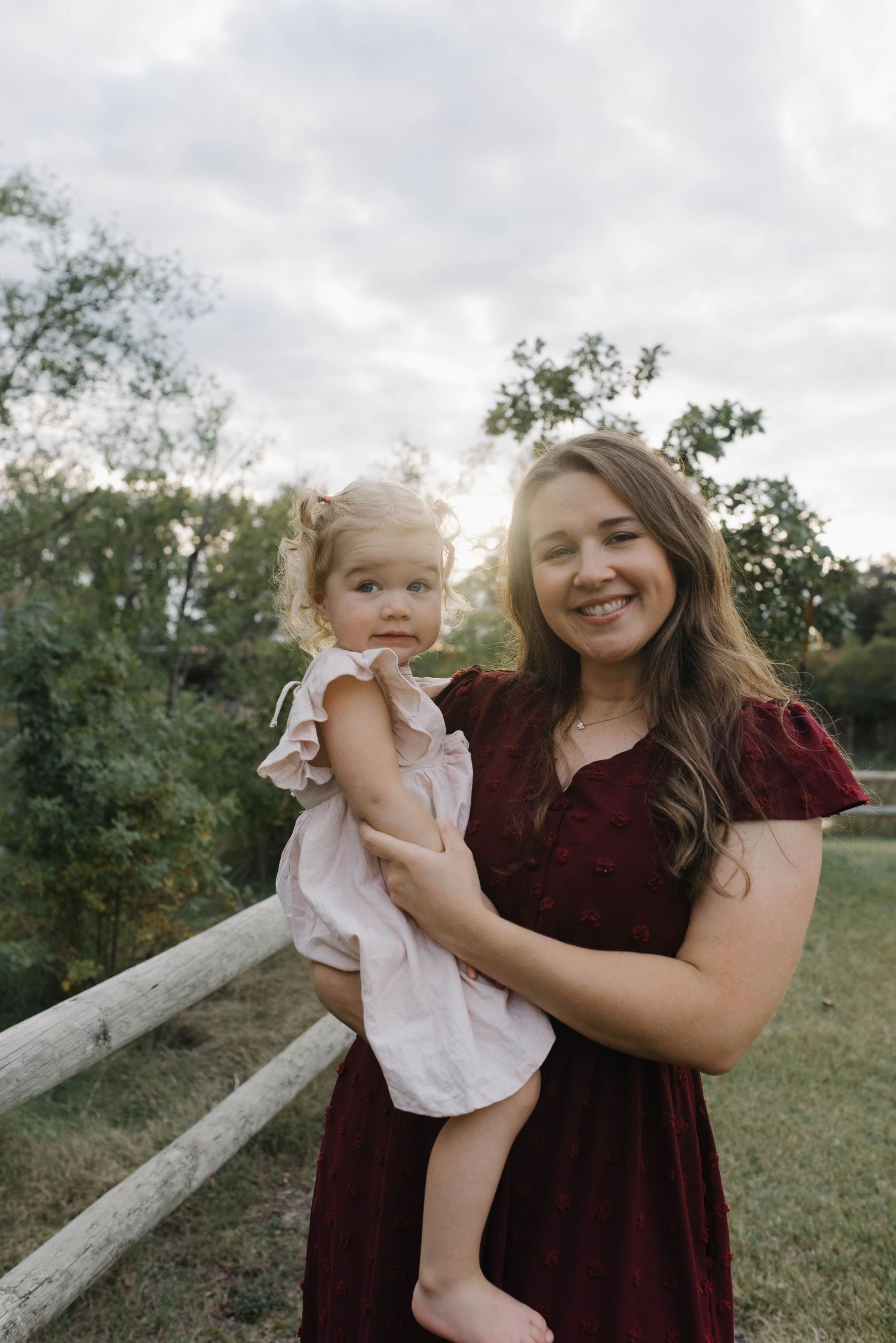 Mother in a burgundy dress smiling while holding her young blonde daughter during an outdoor family photo session.