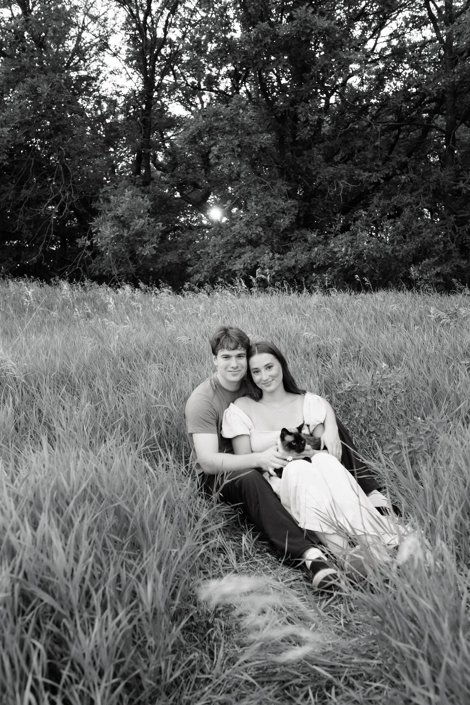 Black and white photo of a couple sitting in a grassy field holding a small cat, with trees in the background.