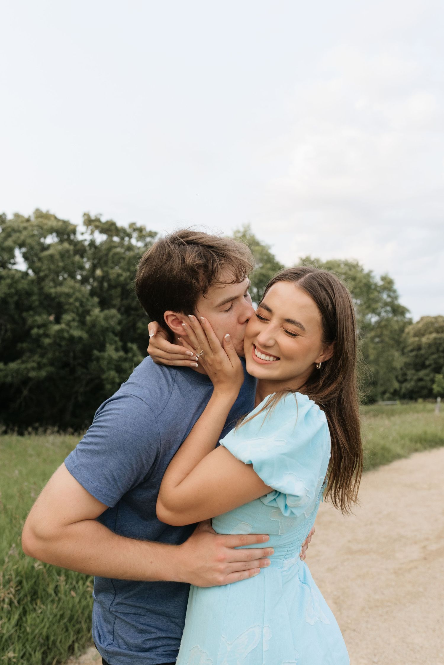 A couple embraces on a nature path, the man kissing the woman's cheek as she smiles joyfully in a light blue dress.