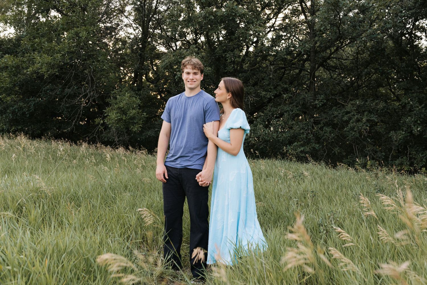 A young couple holds hands in a lush green meadow, the woman in a light blue dress gazing at the man.