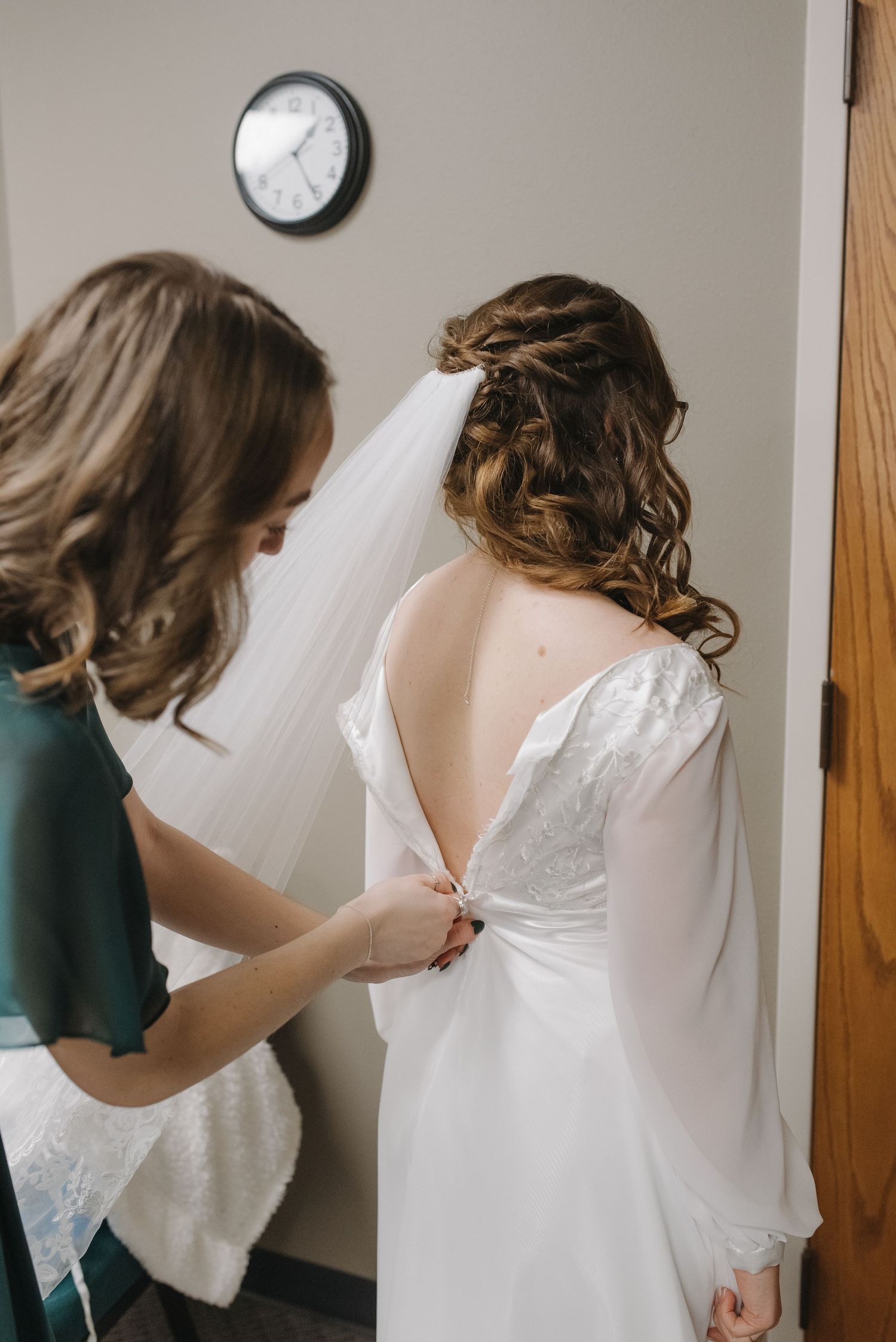 Bridesmaid helping bride button up elegant white backless wedding gown with veil and lace details.