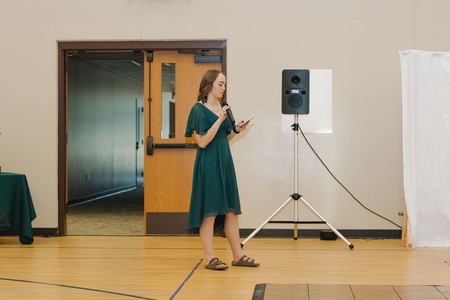 Woman in teal dress speaks at wedding reception podium near speaker on wooden gym floor.
