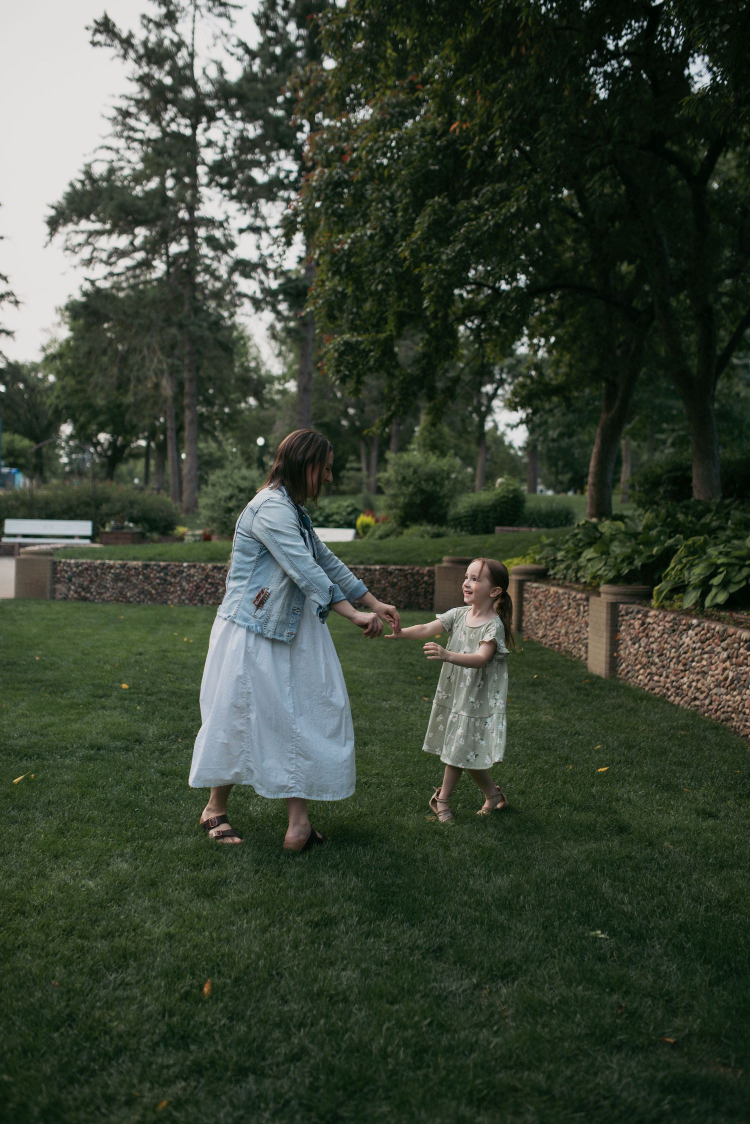 A mother and young daughter hold hands while playing together on green grass in a scenic park setting.