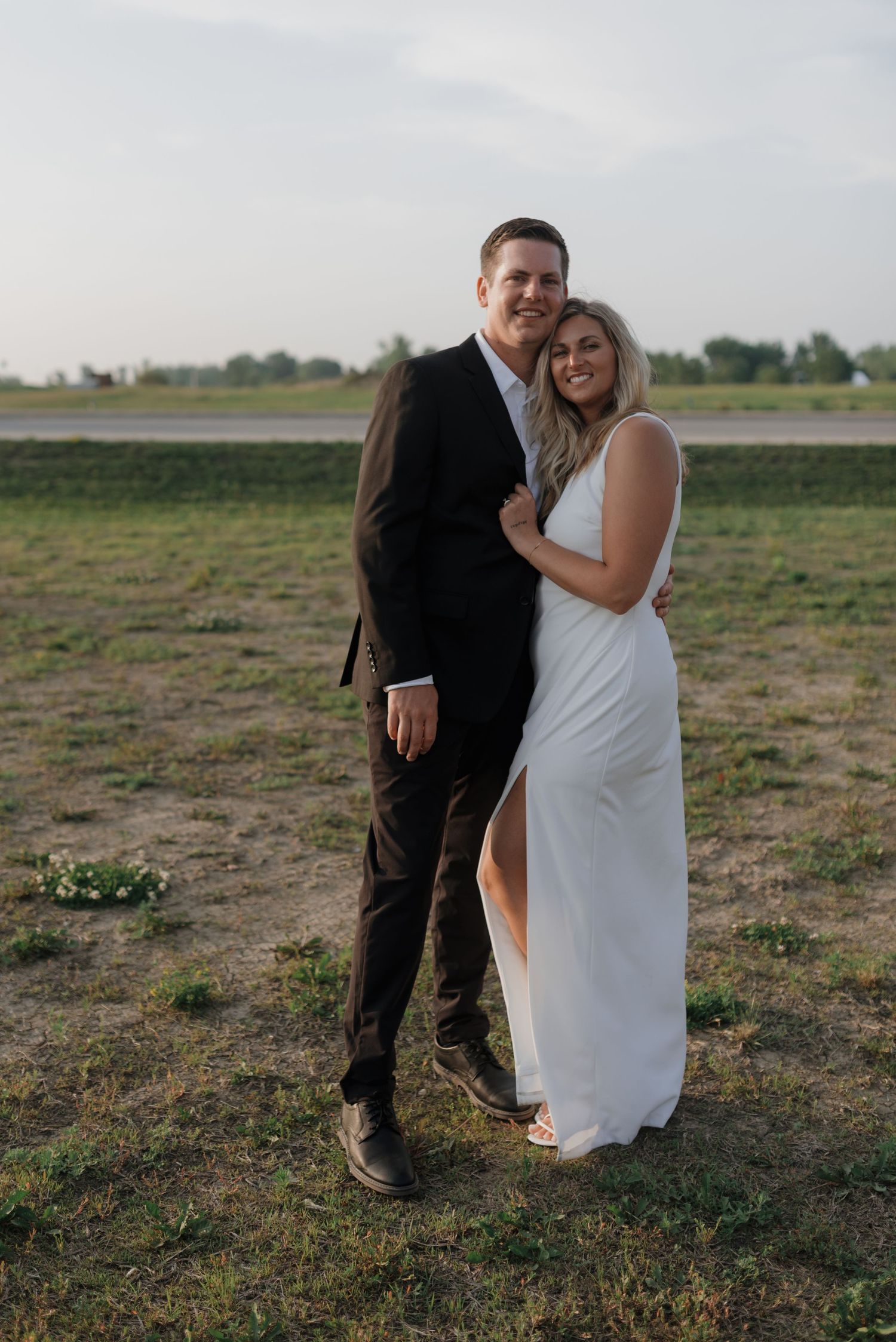 Happy couple embraces in open field at sunset, bride in white slit gown and groom in black suit.