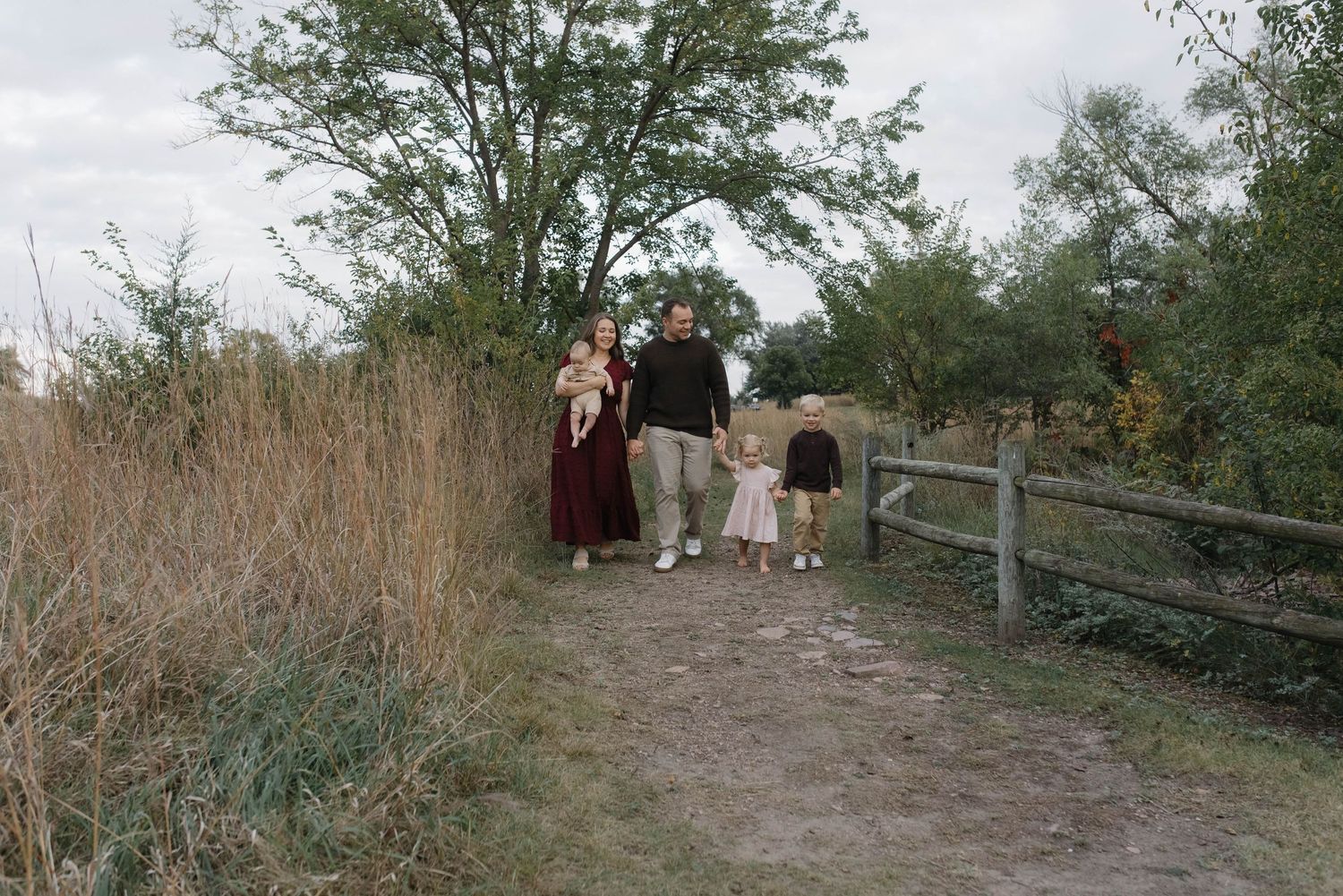 Family of four walking together on dirt path near wooden fence and tall grass outdoors.