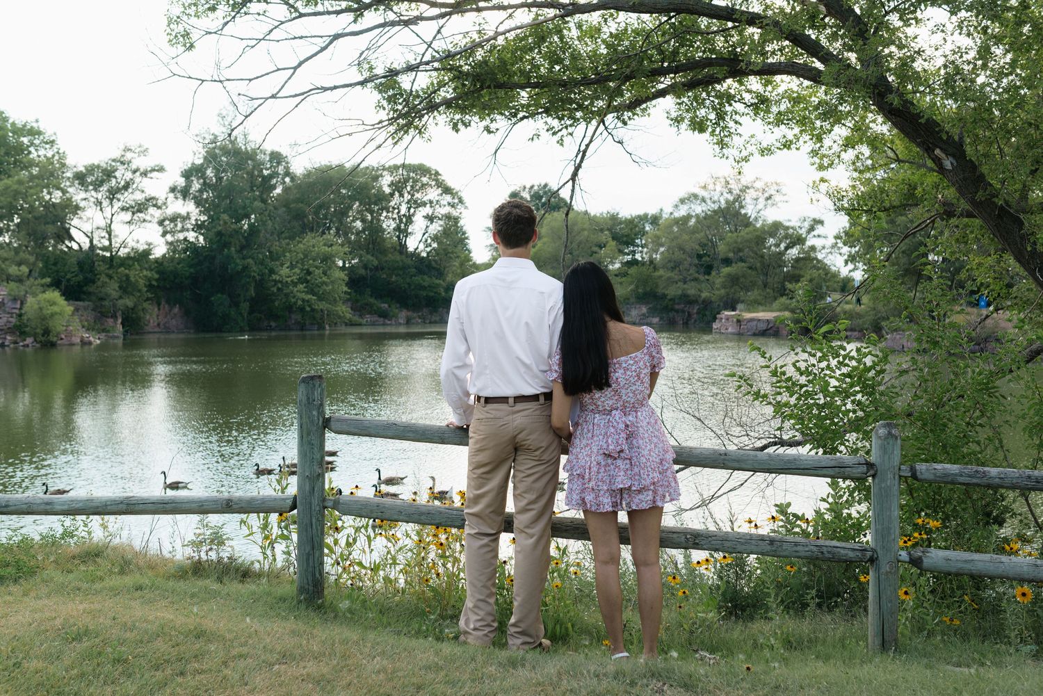 Couple holding hands standing by a wooden fence overlooking a serene pond surrounded by lush green trees.