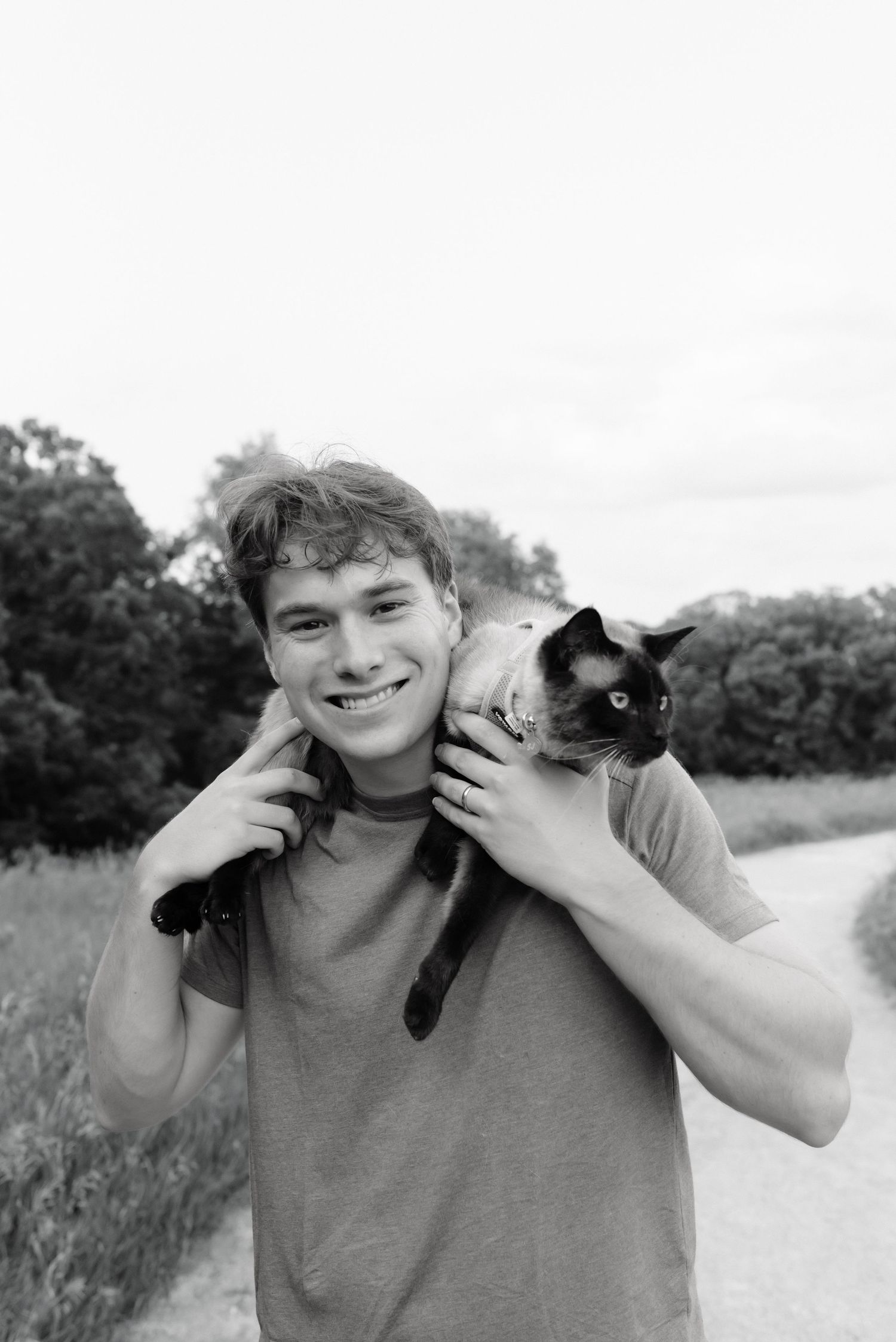 Smiling young man holding a Siamese cat on his shoulders outdoors in a black and white photo.