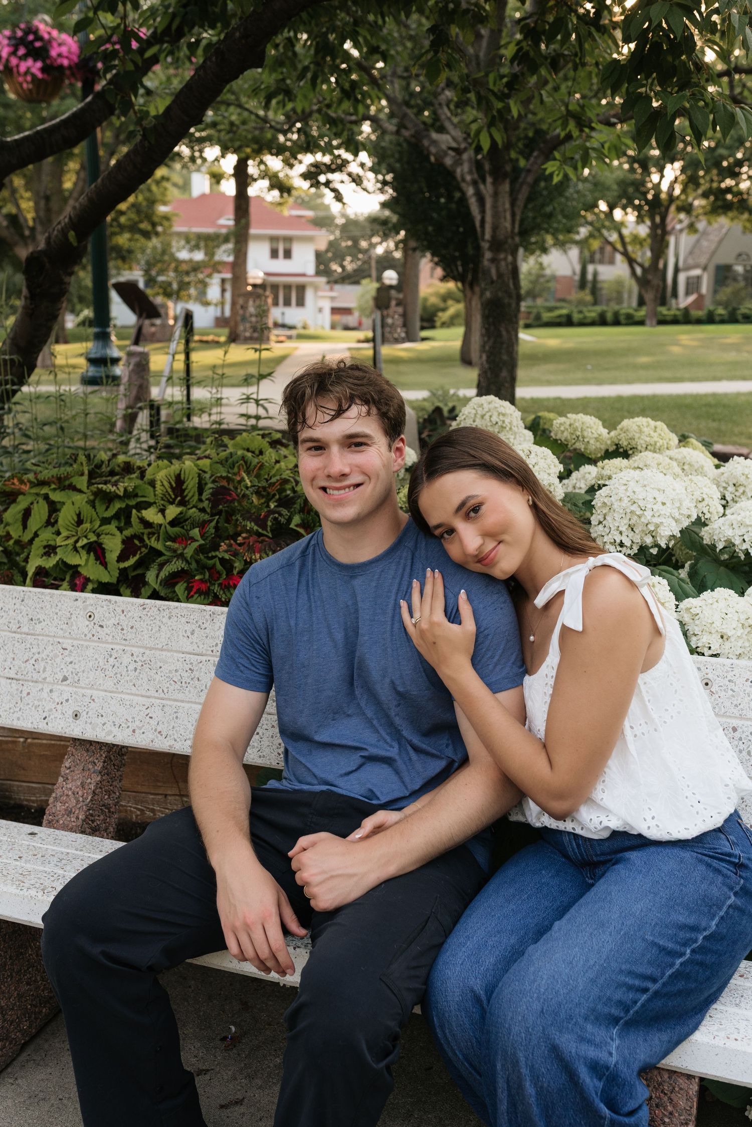 A young couple sits together on a white park bench surrounded by lush greenery and white hydrangeas.