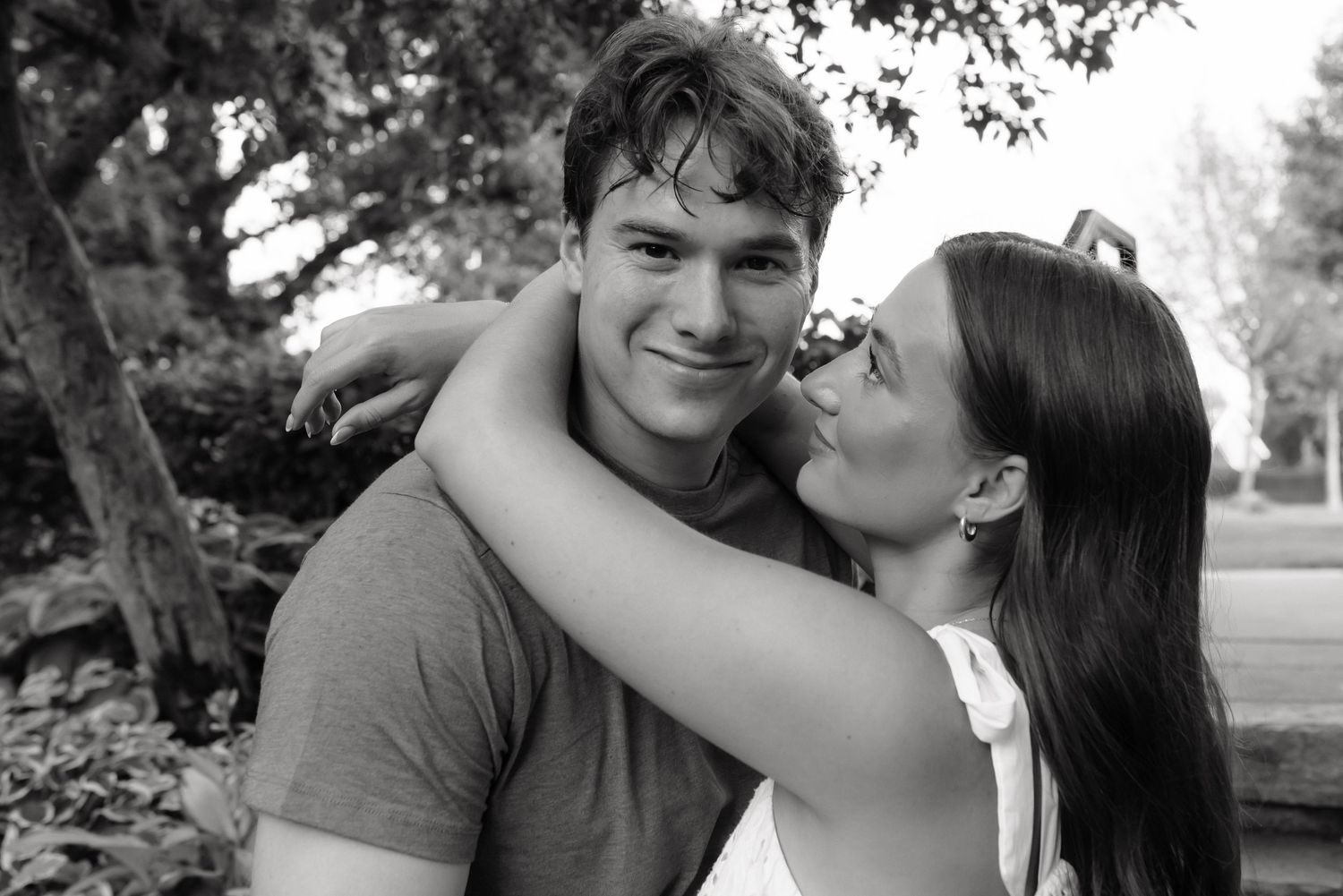 Black and white photo of a smiling young couple outdoors, woman kissing mans cheek while embracing near trees.