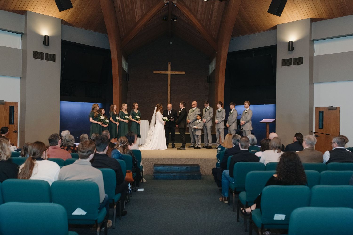 Wedding ceremony in a modern church with bridal party at altar, cross backdrop, and guests seated in teal chairs.