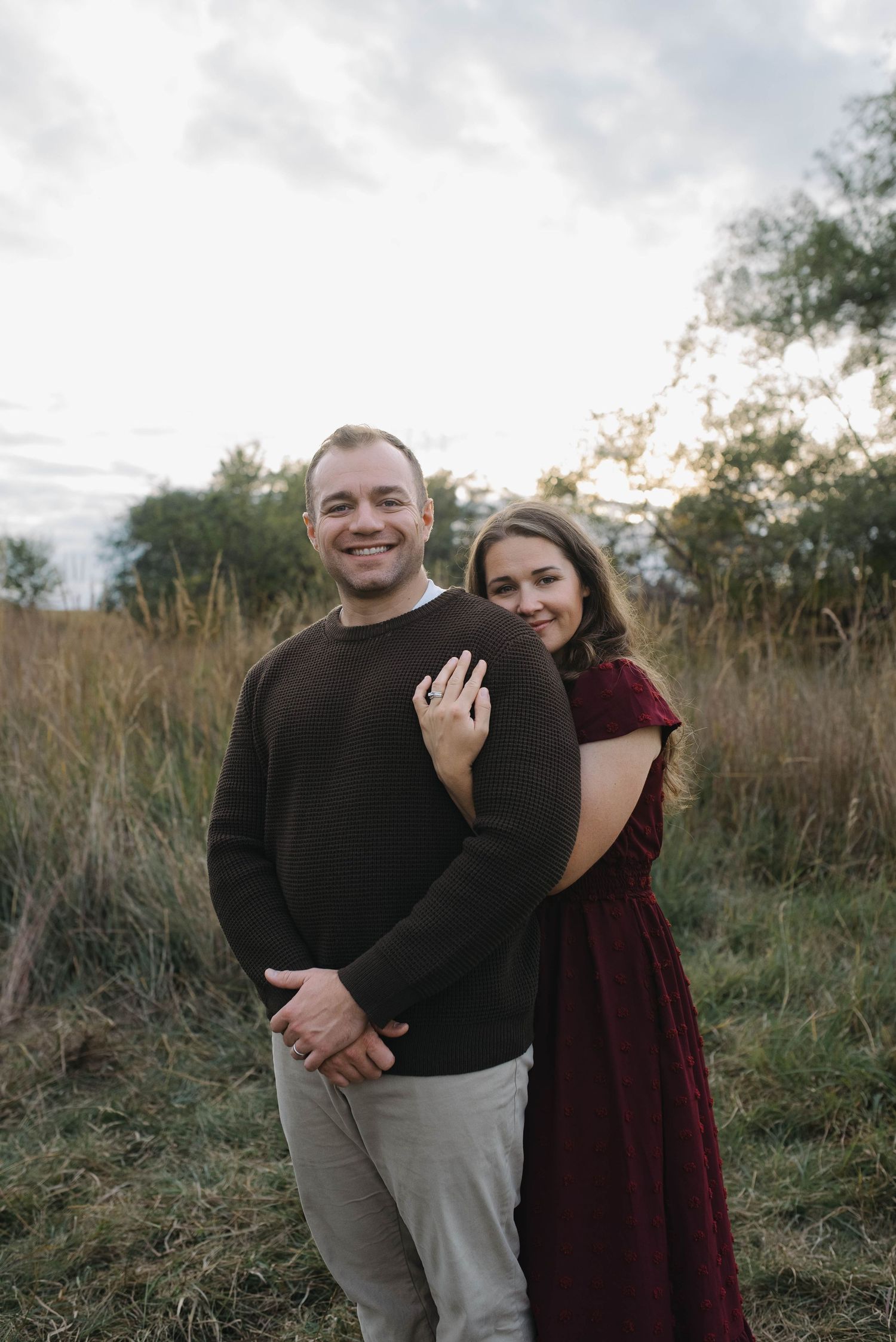 A couple poses in a field, woman in burgundy dress hugging man in dark sweater from behind.