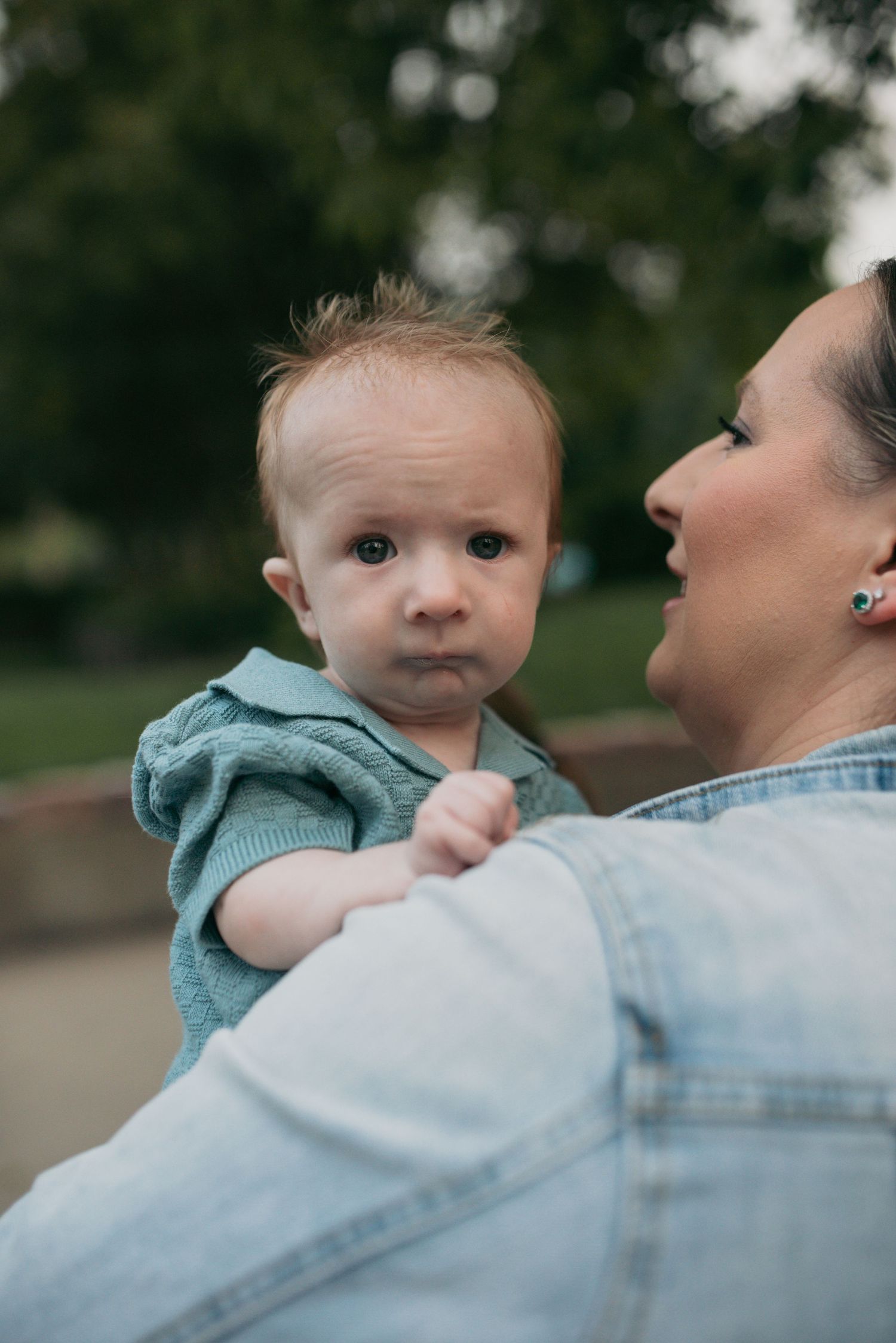 A woman in a denim jacket holding a curious baby dressed in a teal knit romper outdoors.