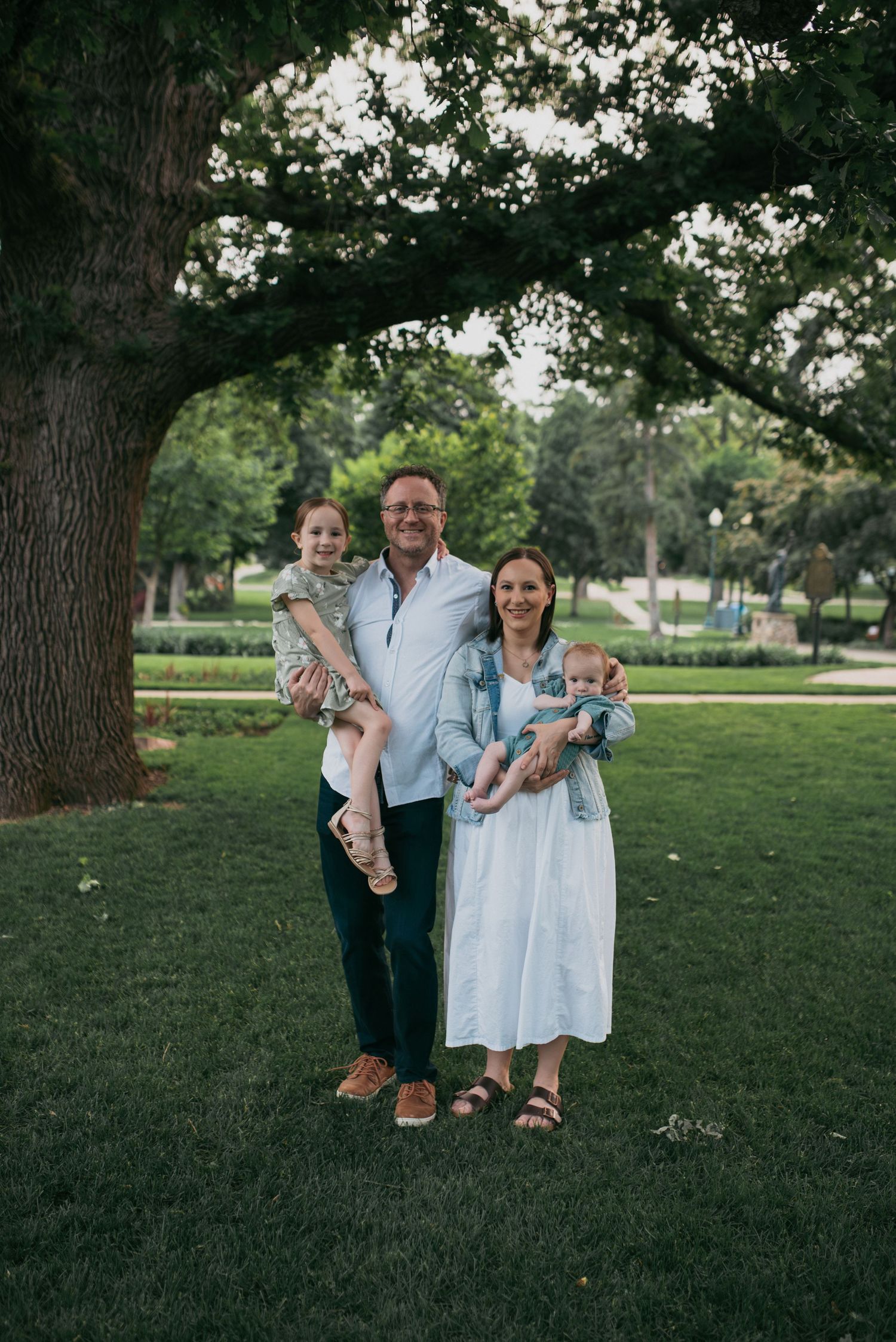 Family of four smiling under a large tree in a park, each parent holding a young child.