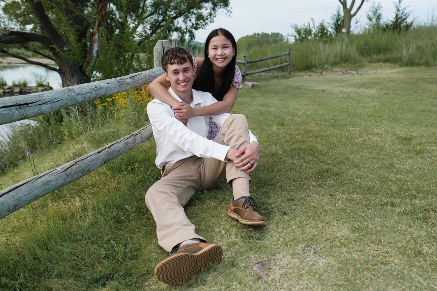 Couple sitting on grass by wooden fence, woman hugging man from behind, smiling outdoors.