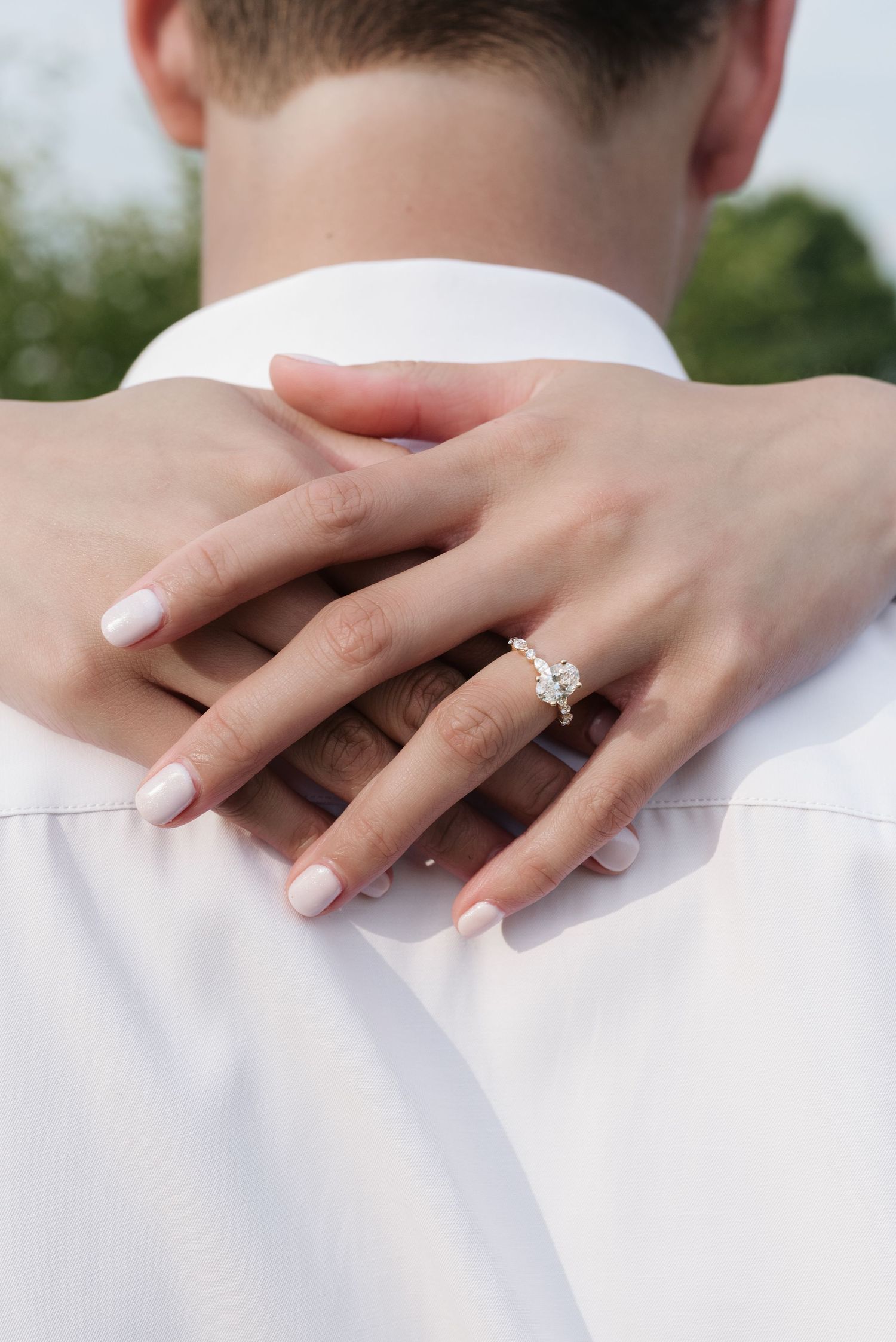 Close-up of a woman's hand with an elegant diamond engagement ring resting on a man's shoulder.