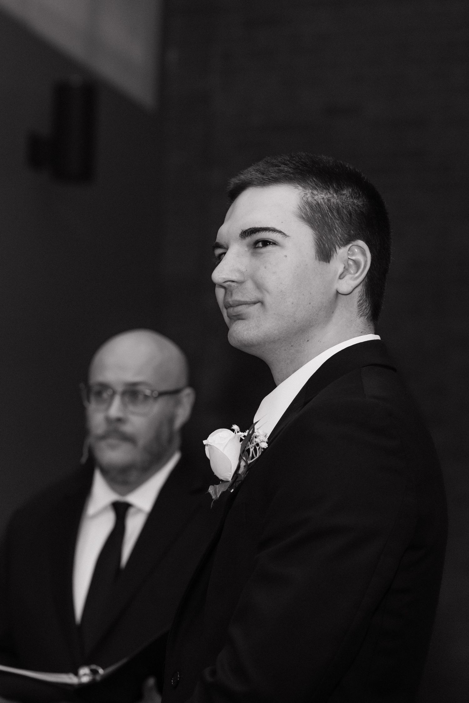 Groom in black suit with white boutonniere smiling at wedding ceremony in black and white photo.