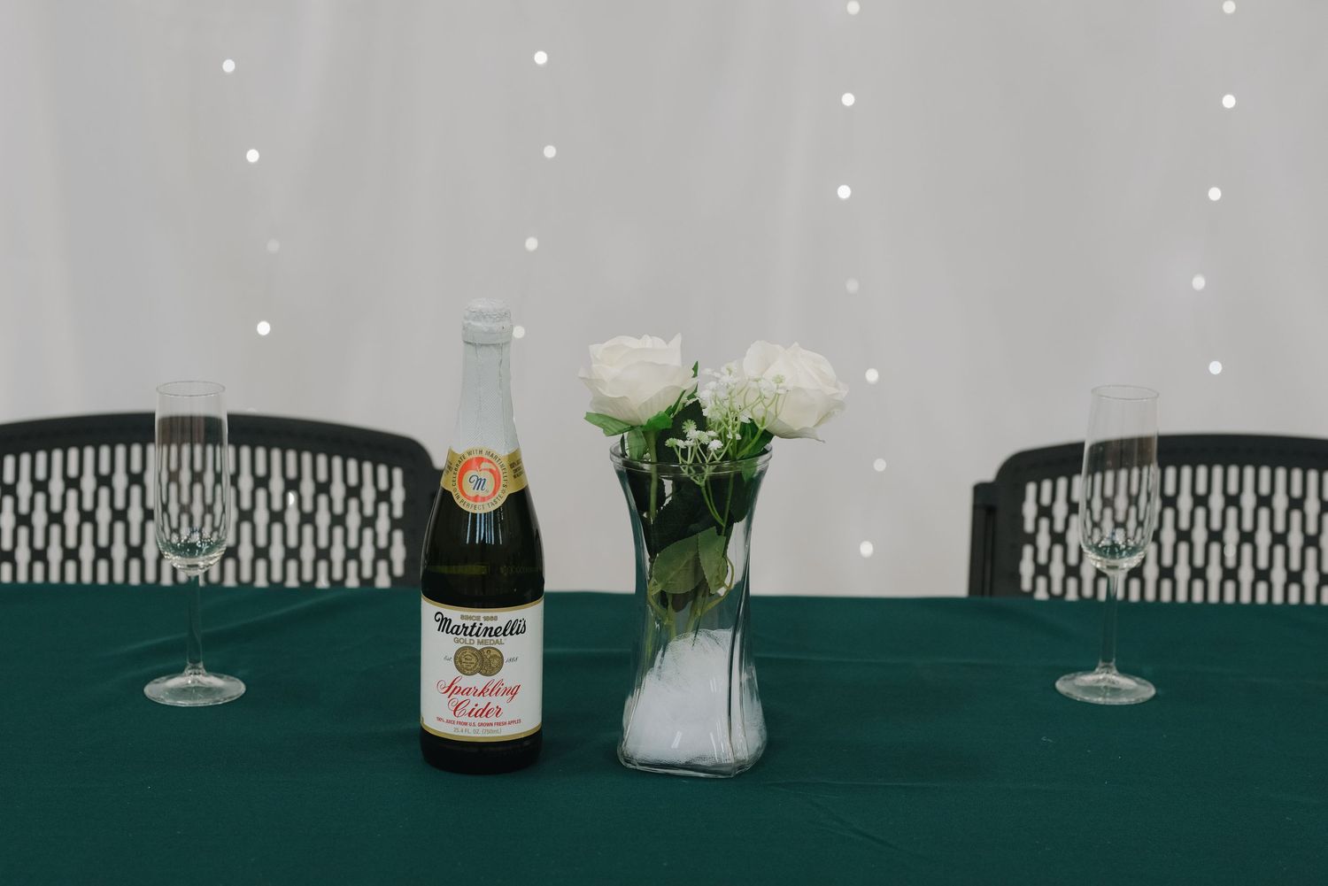 Wedding reception table with sparkling cider bottle, white roses in vase, and champagne glasses.